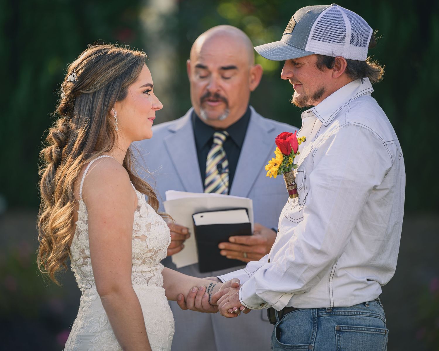 Bride and groom holding hands during outdoor wedding ceremony with officiant reading from a book.