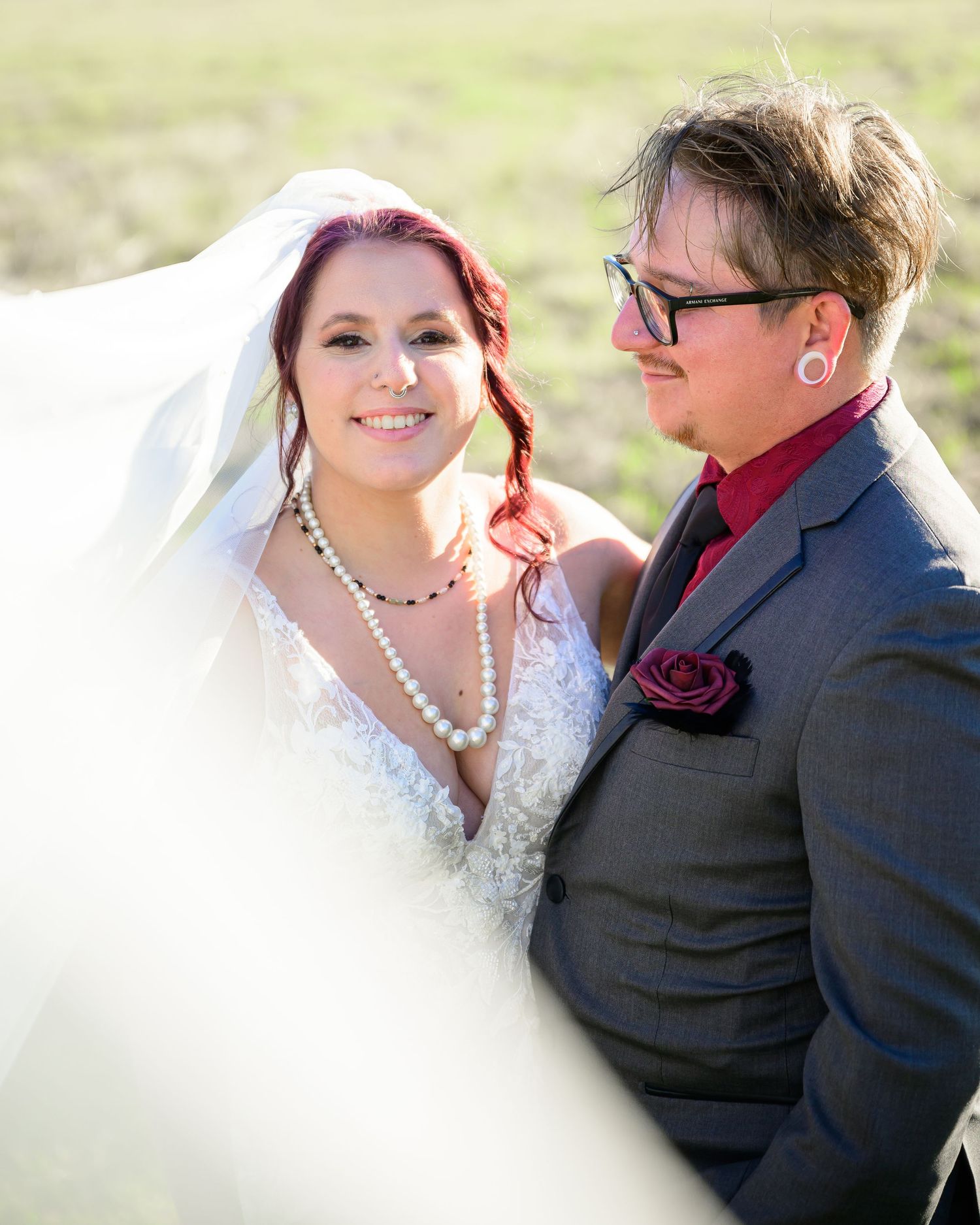 Smiling bride in lace gown and pearl necklace with groom in gray suit at outdoor wedding.