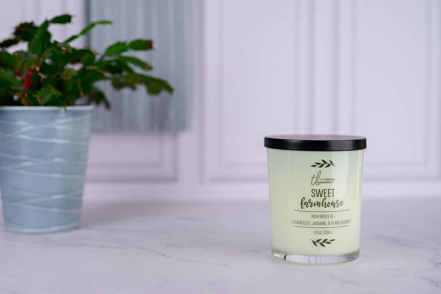 A Sweet Farmhouse scented candle in a glass jar displayed on a white surface beside a green houseplant.