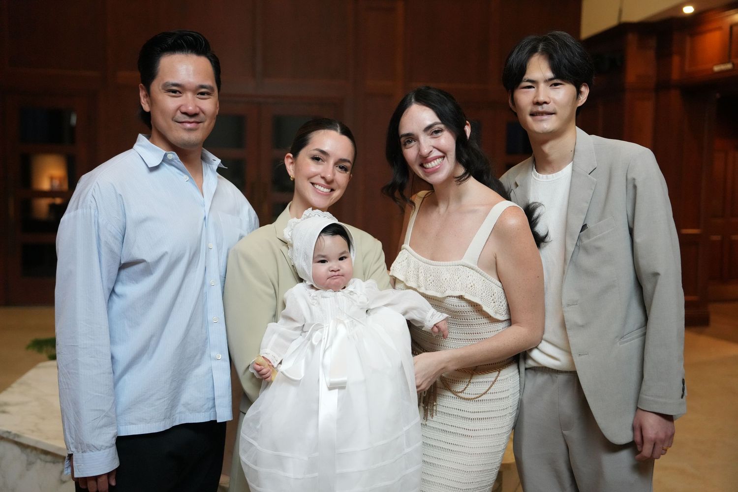Family gathered for a baby baptism ceremony, with infant in white christening gown held by woman in white dress.