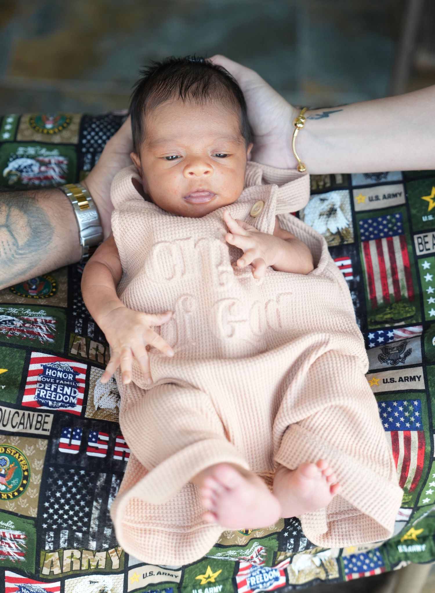 Newborn baby in pink onesie rests on colorful US Army patchwork blanket.