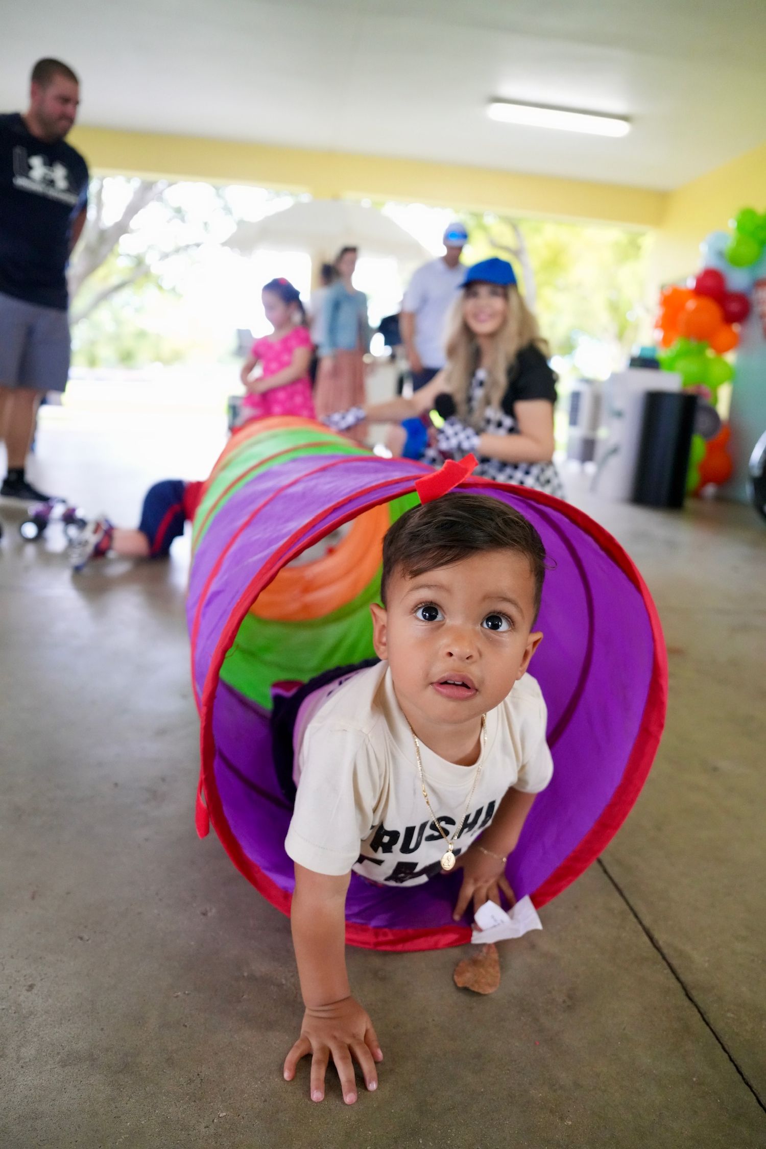 Curious toddler crawls through colorful play tunnel at outdoor birthday party.