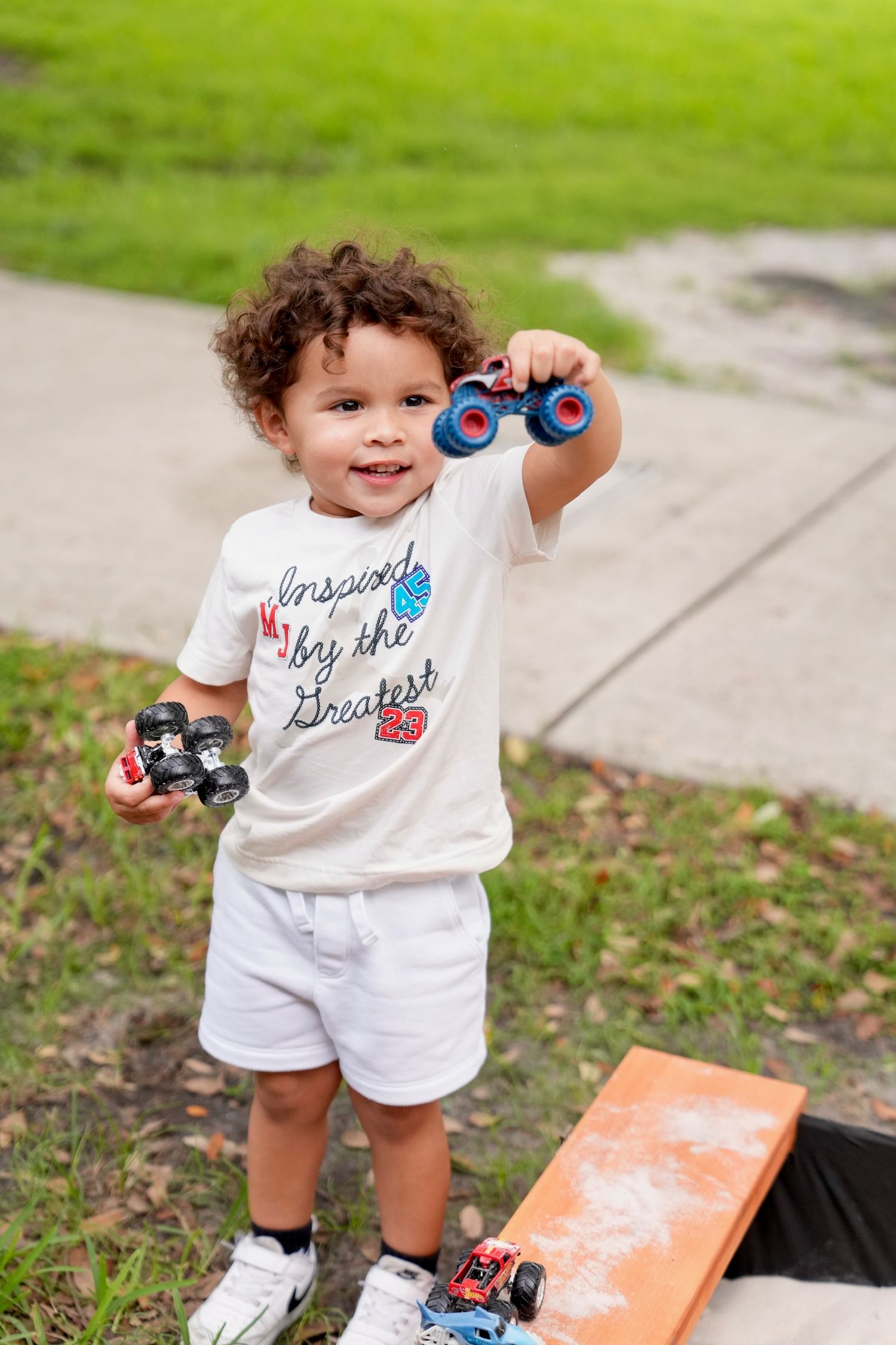 A curly-haired toddler smiles while holding toy monster trucks outdoors.
