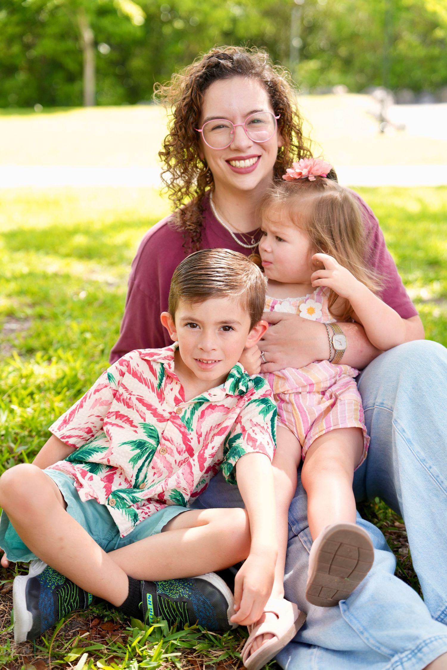 A smiling woman with curly hair and glasses sits on grass with two young children in a sunny park.