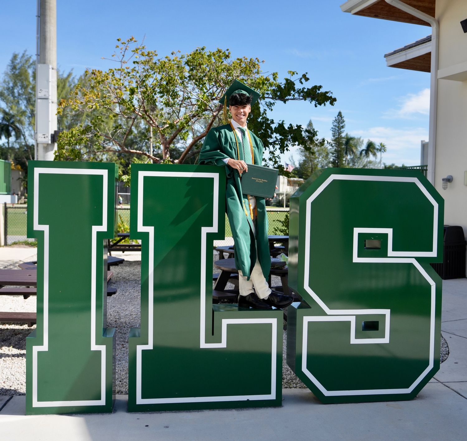 A graduate in green cap and gown poses on large ILS school letter decorations.