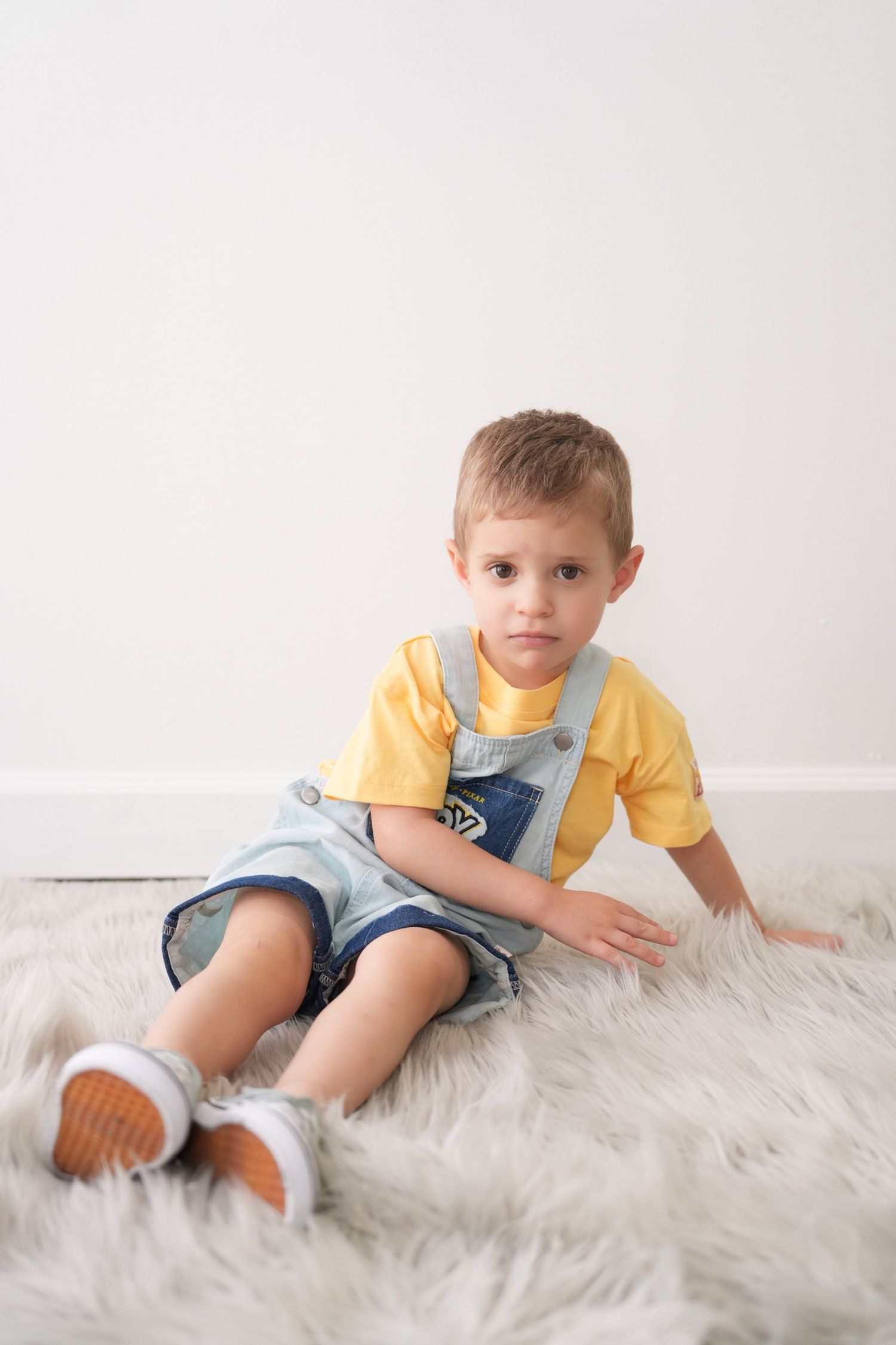 A young boy in denim overalls and yellow shirt sits on a fluffy white rug.