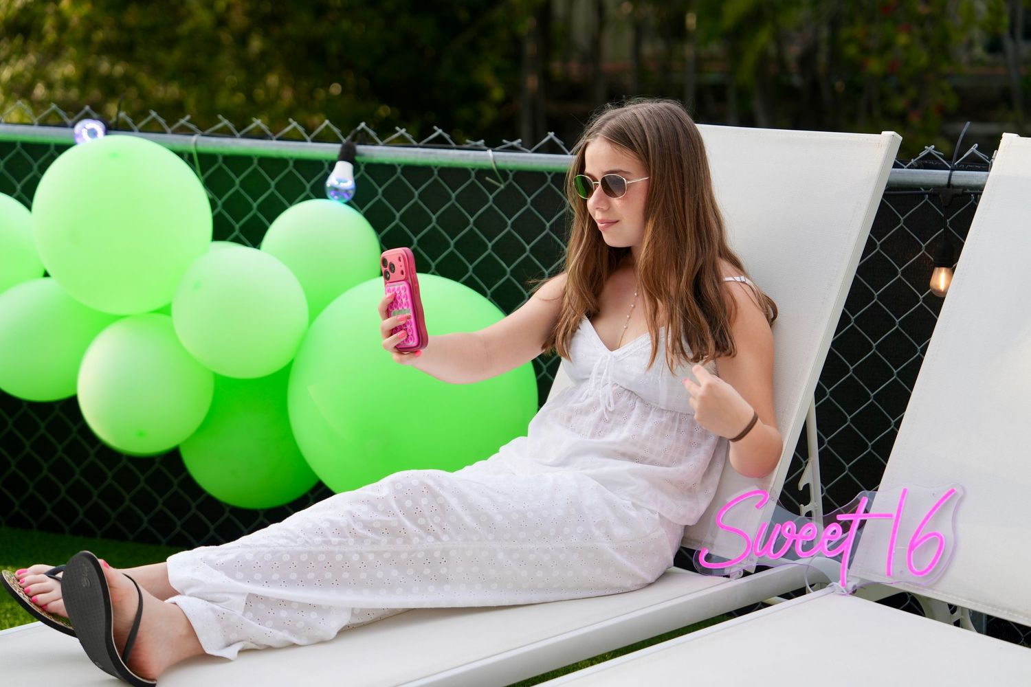 A teenage girl in white outfit takes a selfie by green balloons at a Sweet 16.