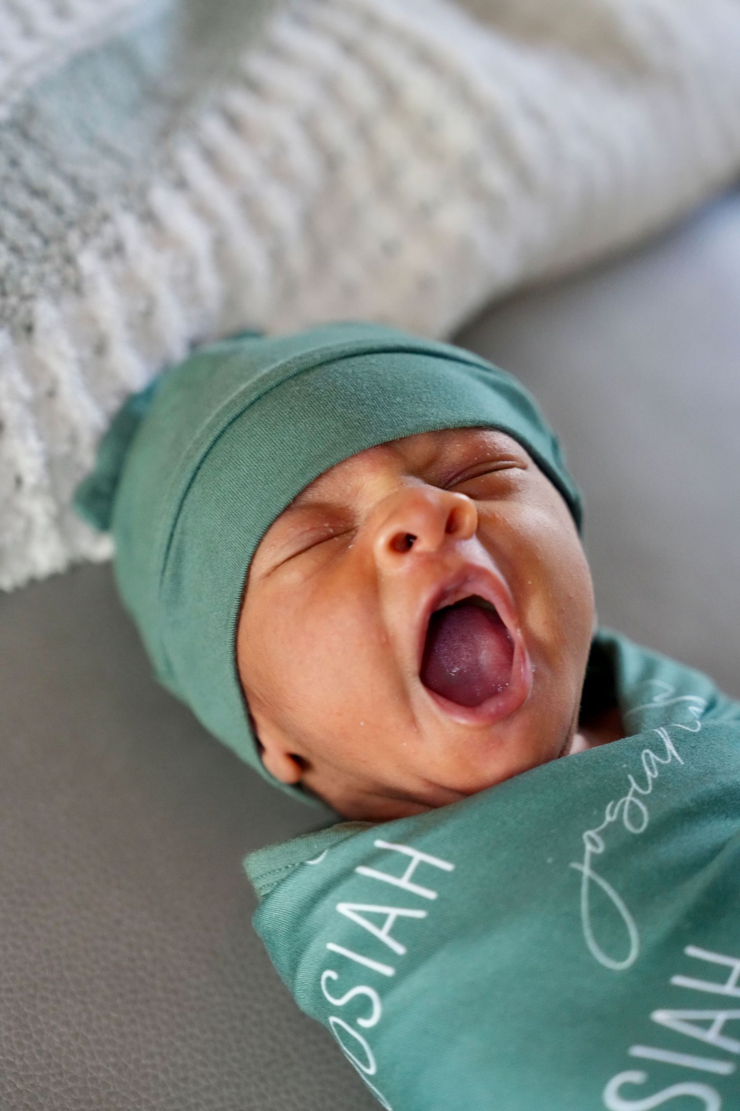 A newborn baby named Josiah yawns while wrapped in a teal personalized swaddle.