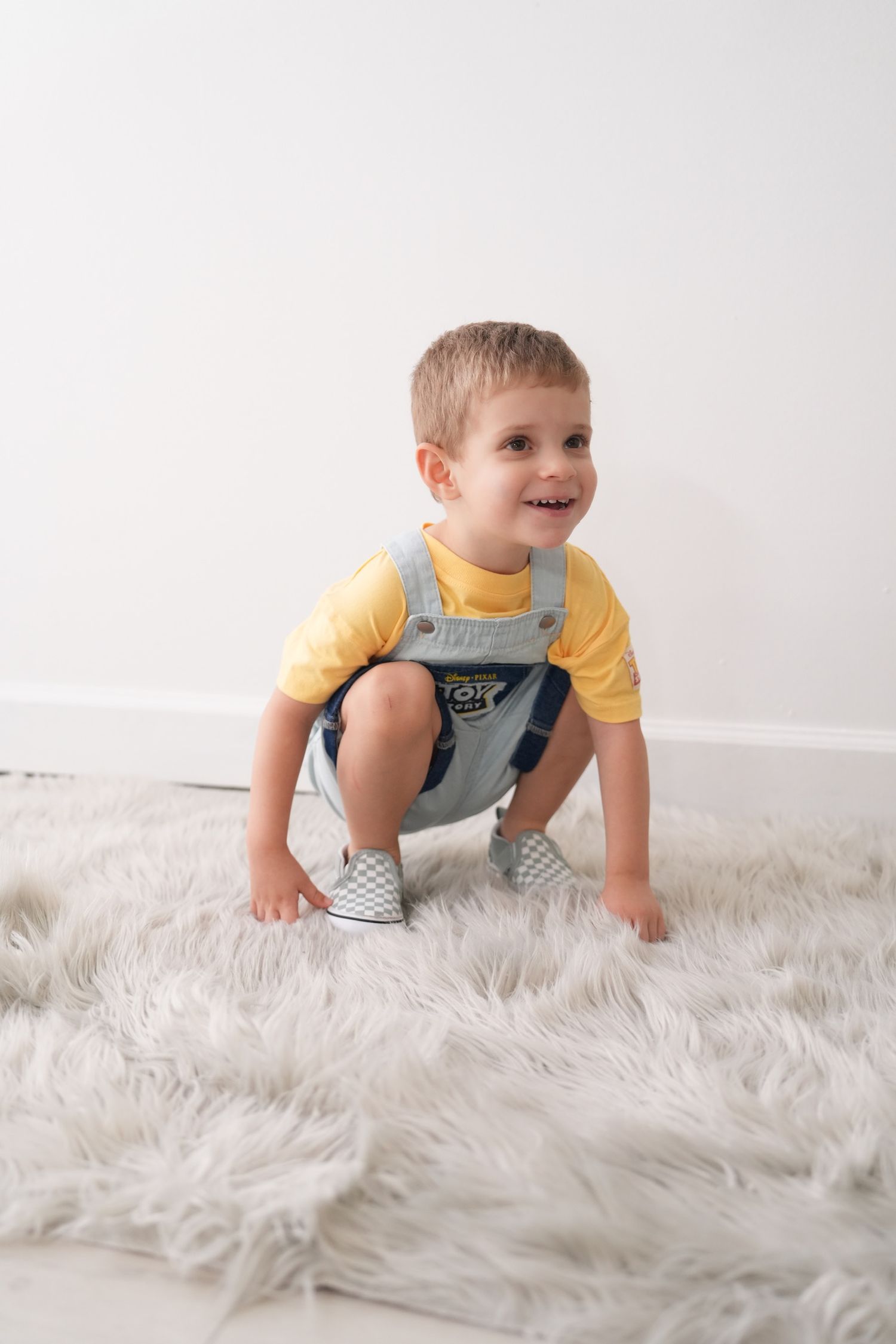 Happy toddler in yellow shirt and denim overalls crouches on fluffy white rug.