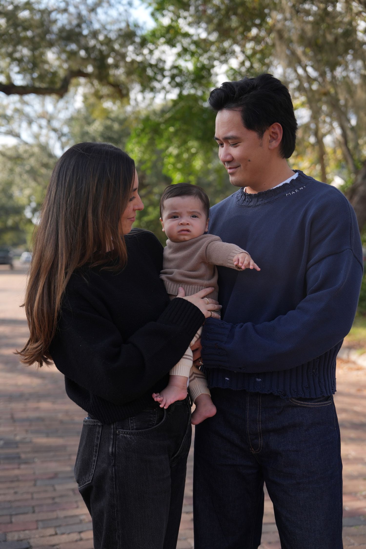 Young family with baby poses outdoors on brick path surrounded by trees.