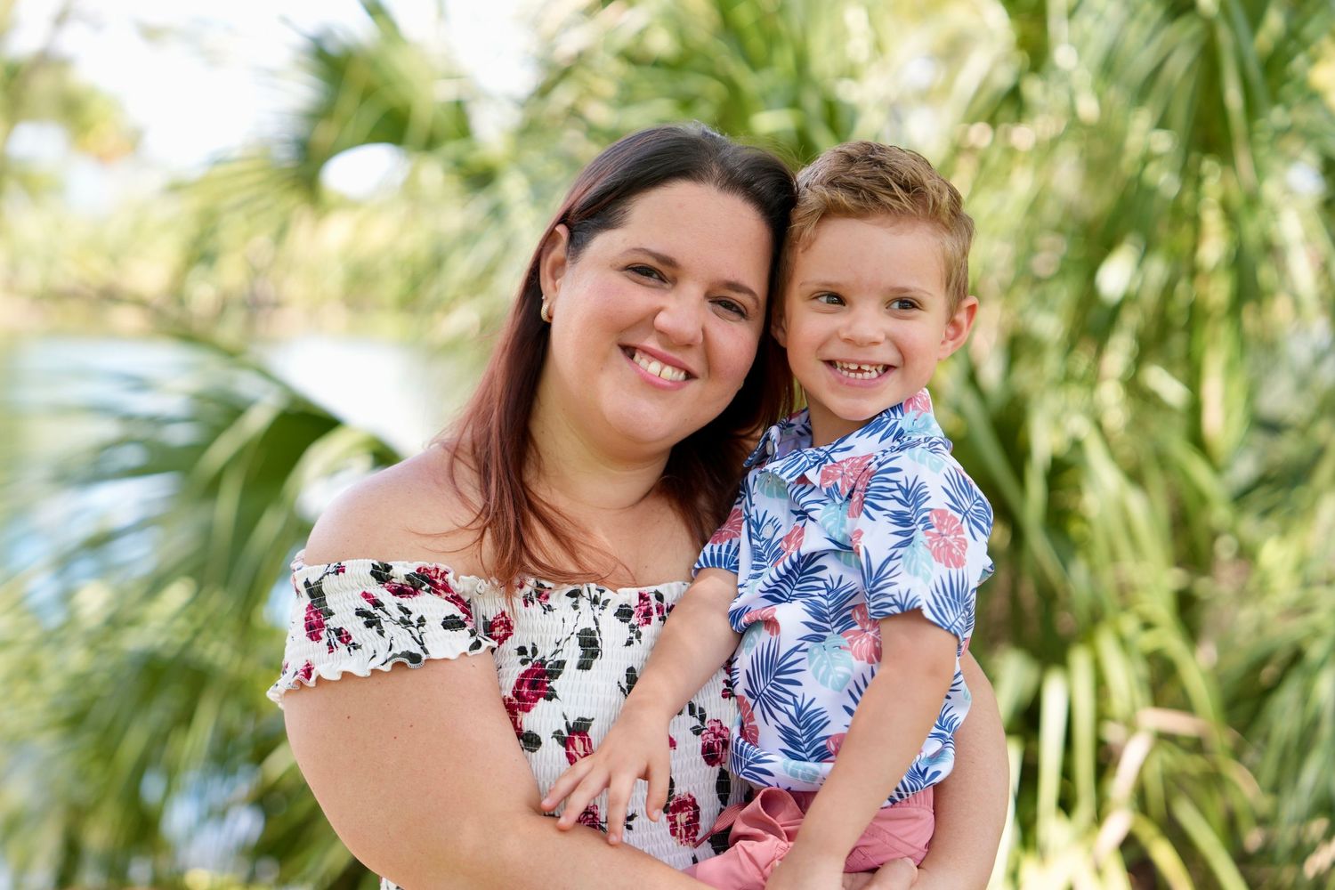 A smiling woman holds a young boy in a floral shirt outdoors near palm trees and a serene water backdrop.