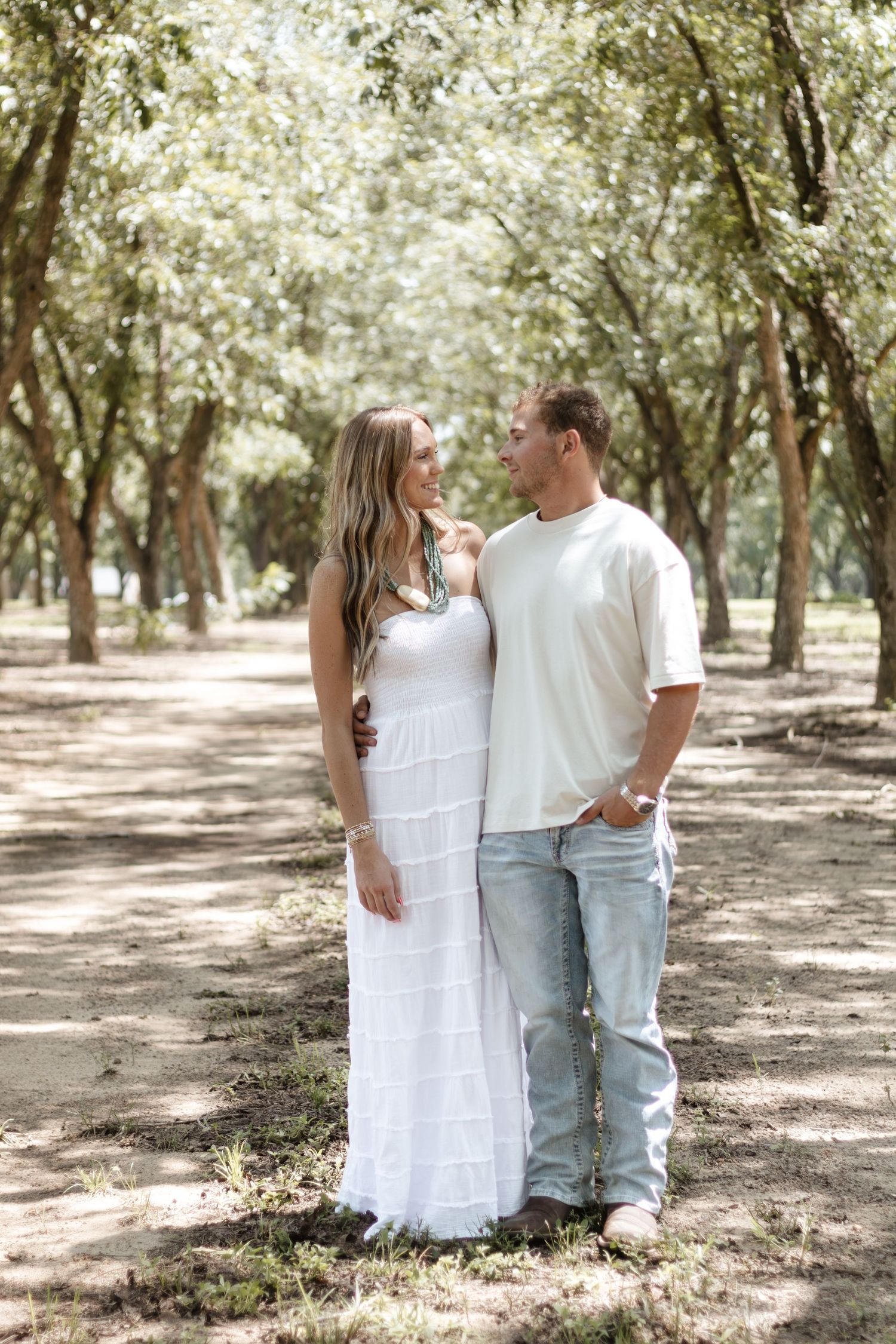 Couple in white outfits gazing at each other lovingly while standing on a shaded tree-lined path.