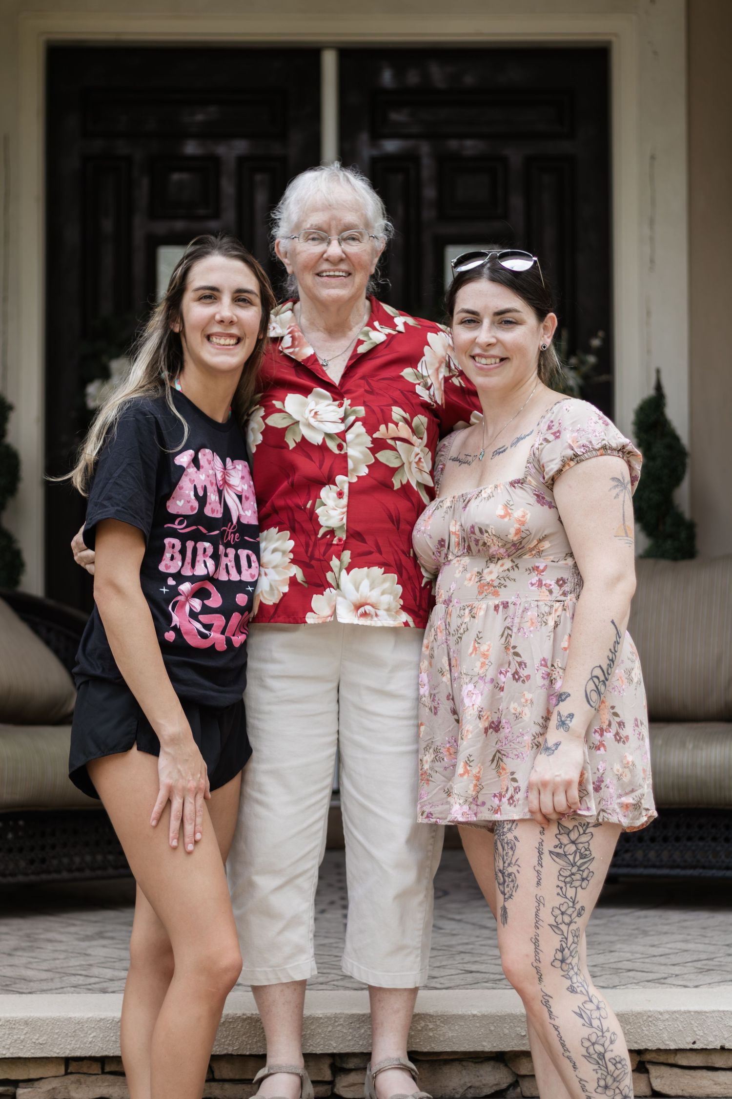 Three women of different generations smile together on a home's front porch steps outdoors.