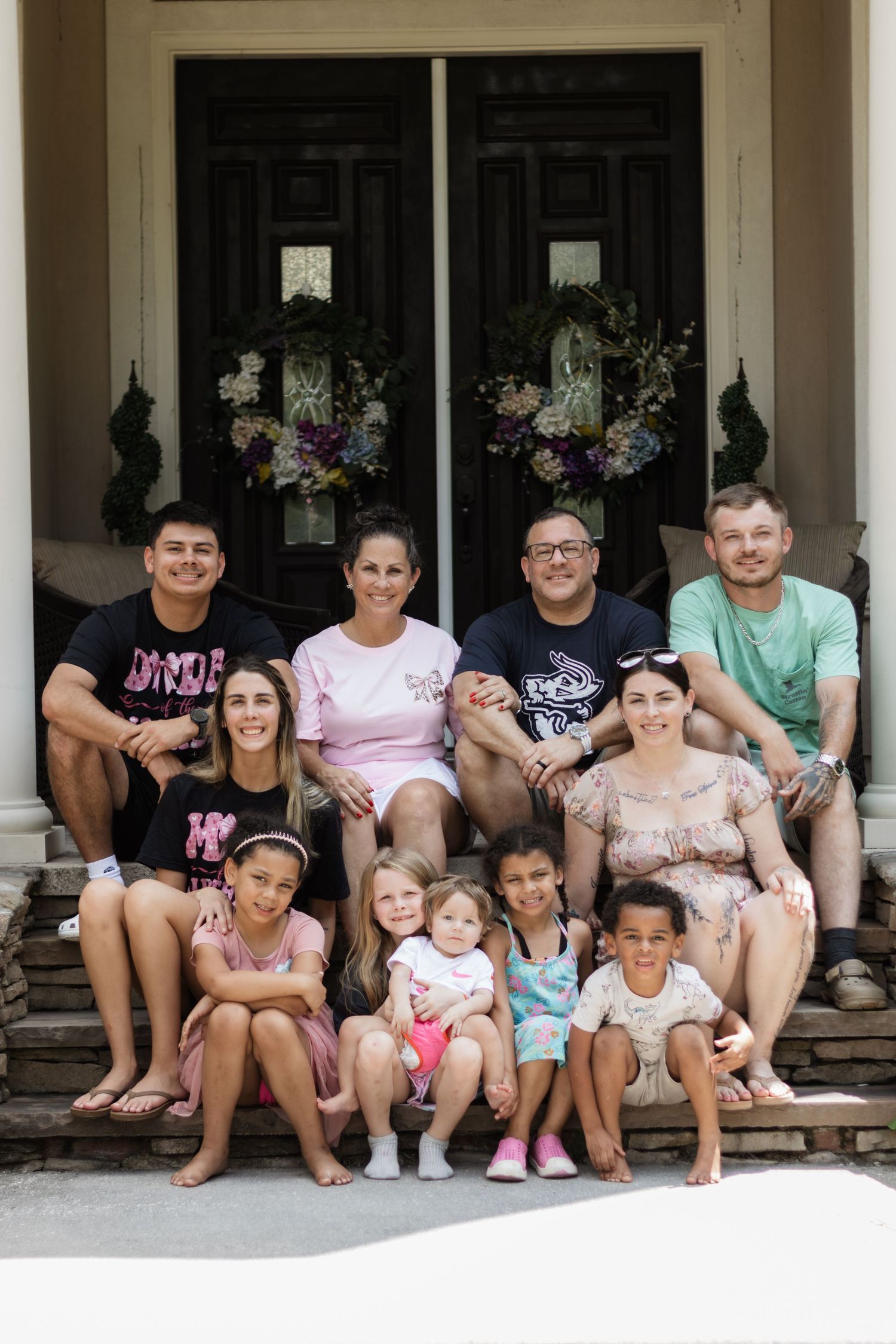 A large multigenerational family poses together on front porch steps outside a home with floral wreaths.