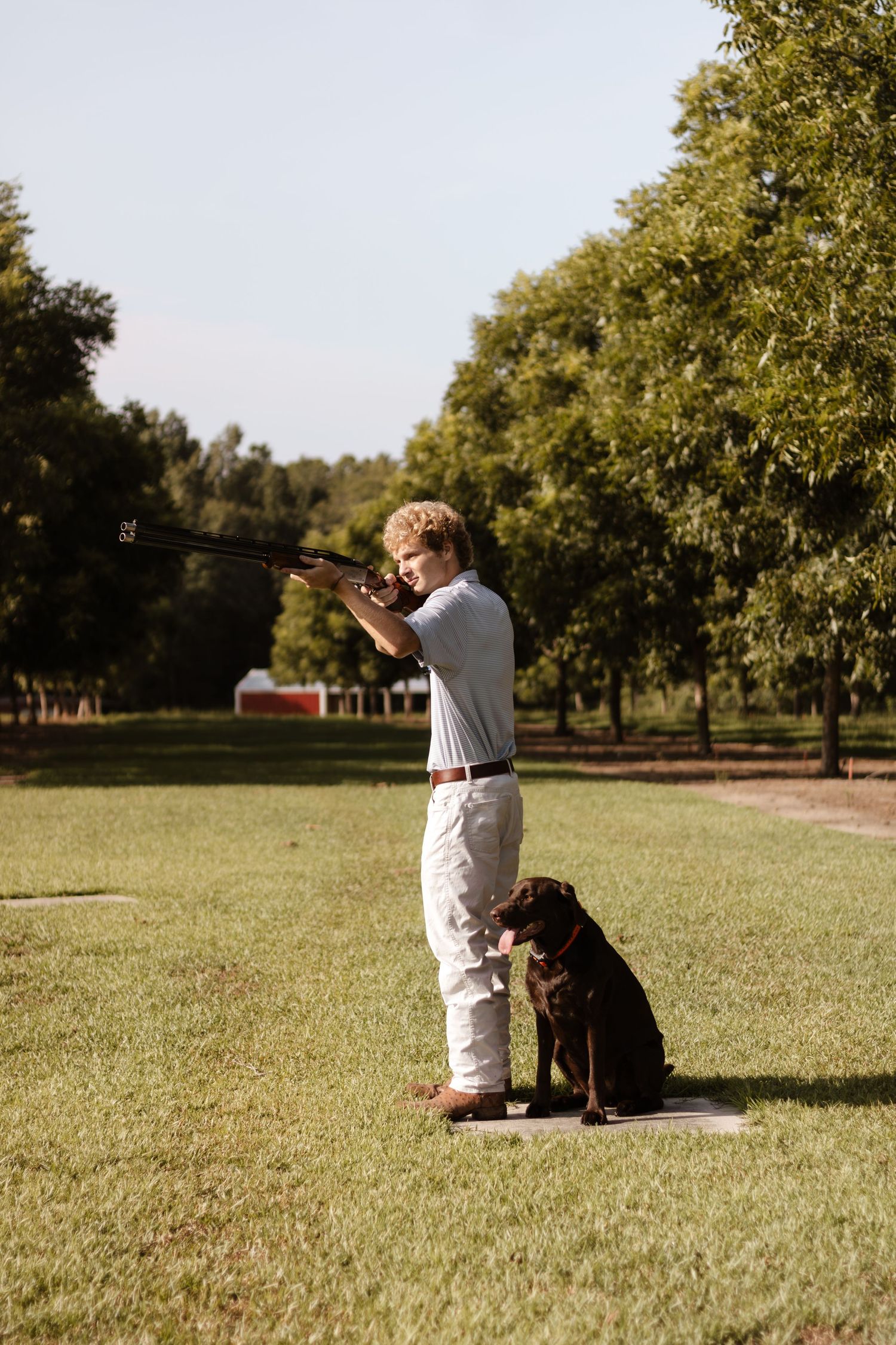 Young man aiming a shotgun while a chocolate Labrador Retriever sits beside him on a green lawn.