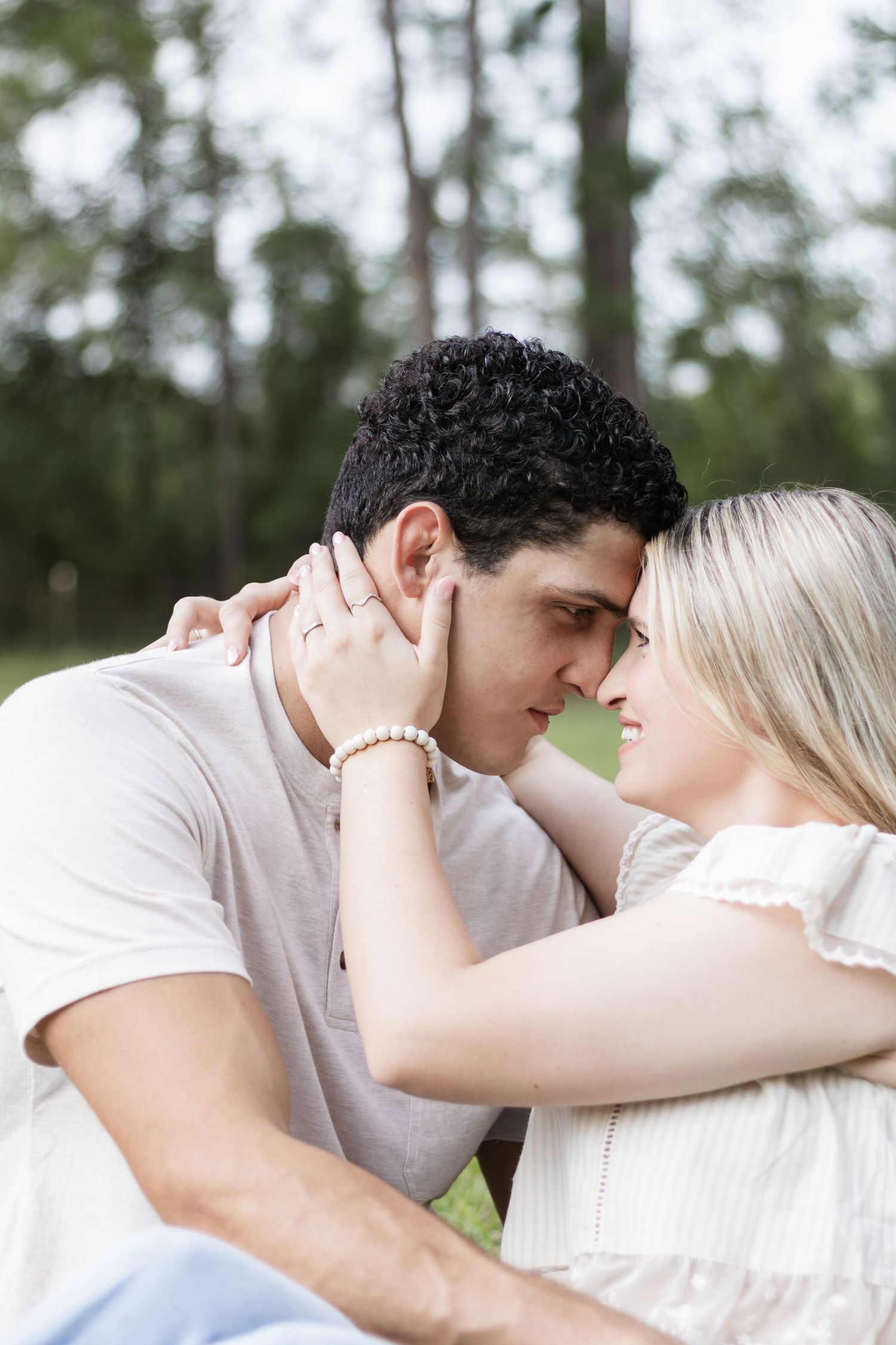 A young couple gazes into each other's eyes while sitting close together outdoors among pine trees.