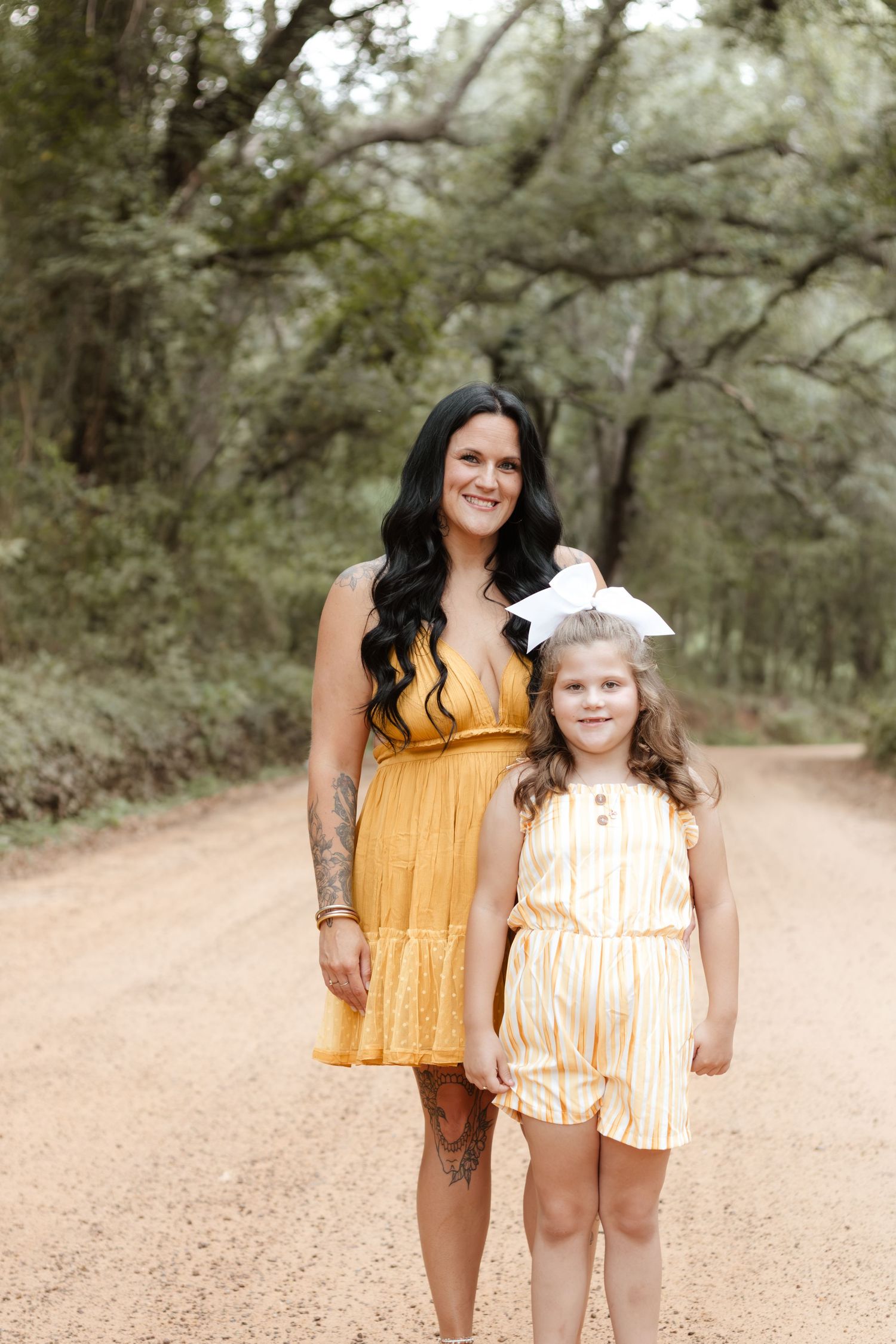 A mother in a mustard yellow dress and her daughter in a yellow striped romper pose on a tree-lined dirt path.