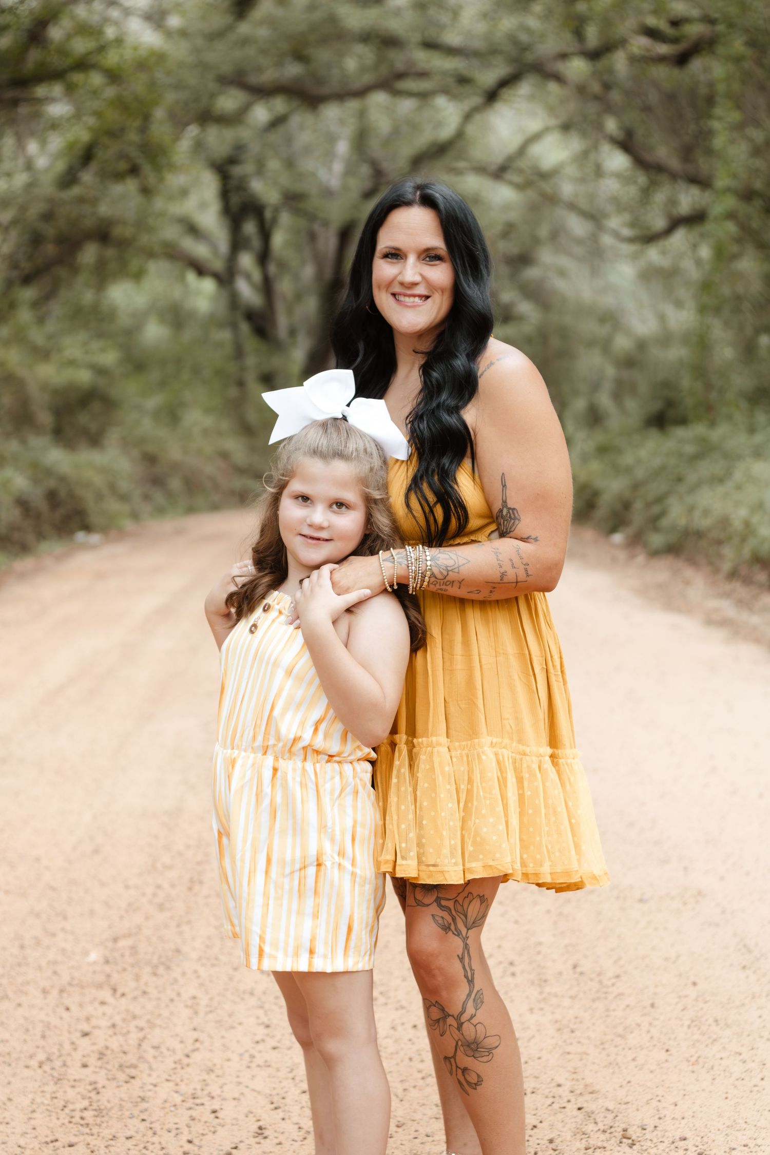 A smiling woman in a yellow dress hugs a young girl in a striped romper with a white bow on a dirt road.