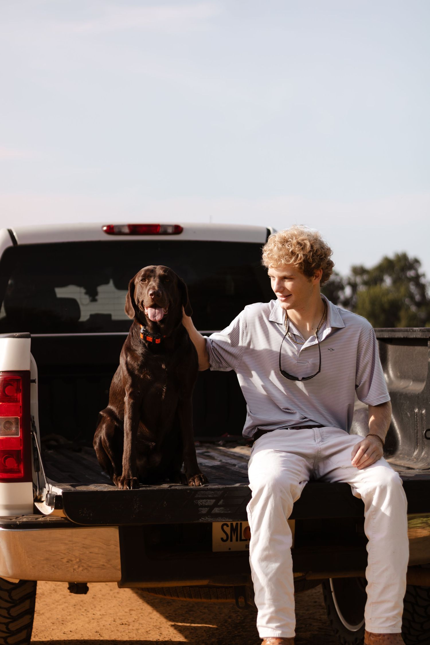 Young man with curly hair sitting on truck tailgate petting a chocolate Labrador Retriever outdoors.