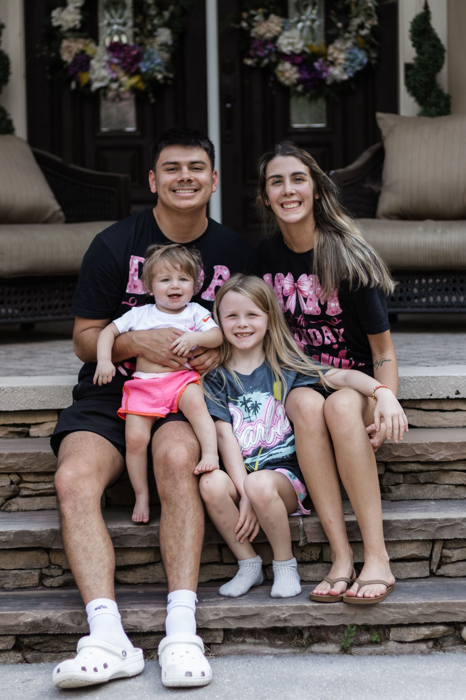 A young family of four smiles together on stone steps in front of a decorated front door.