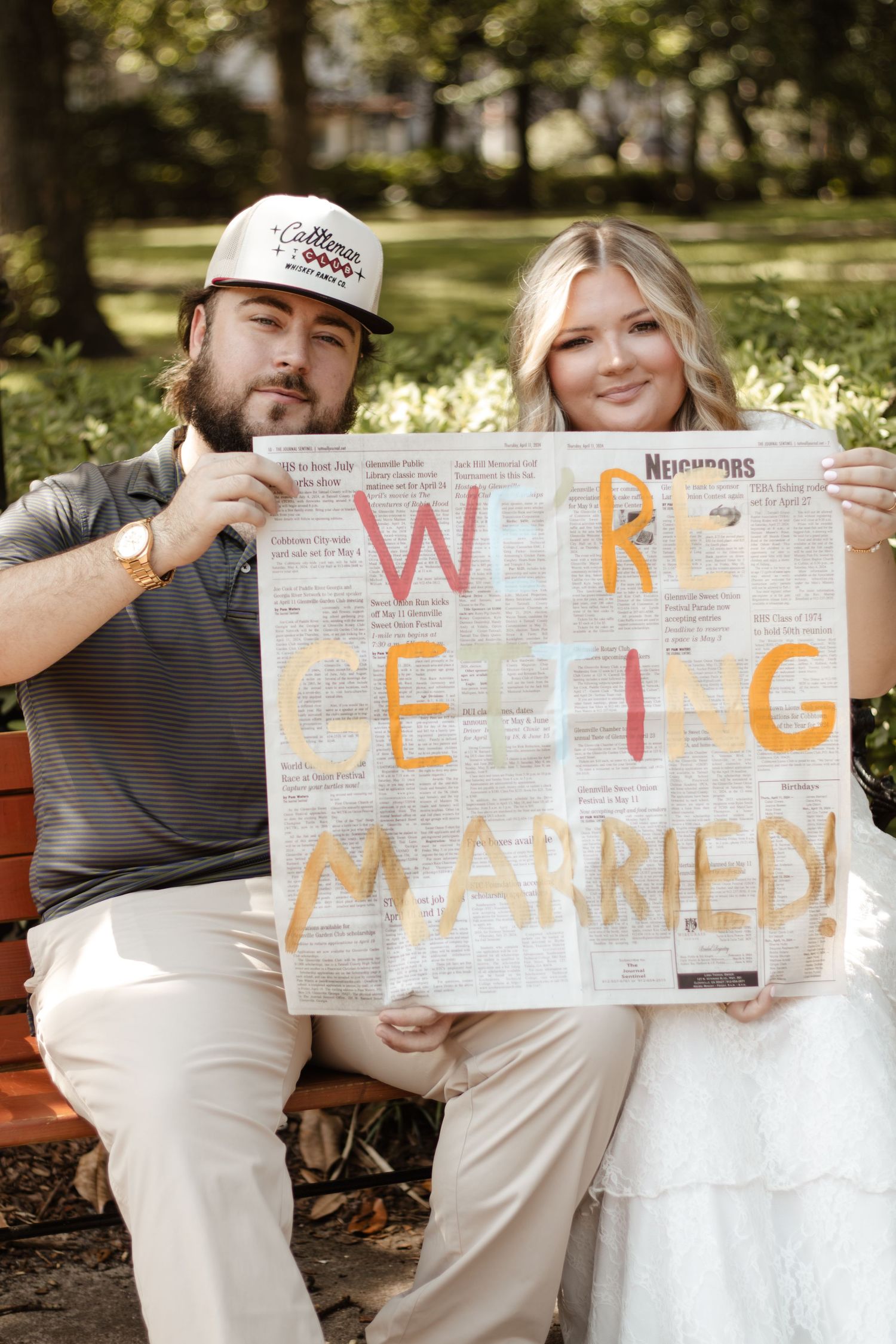 Couple holding a newspaper sign reading We're Getting Married for a fun engagement announcement photo outdoors.