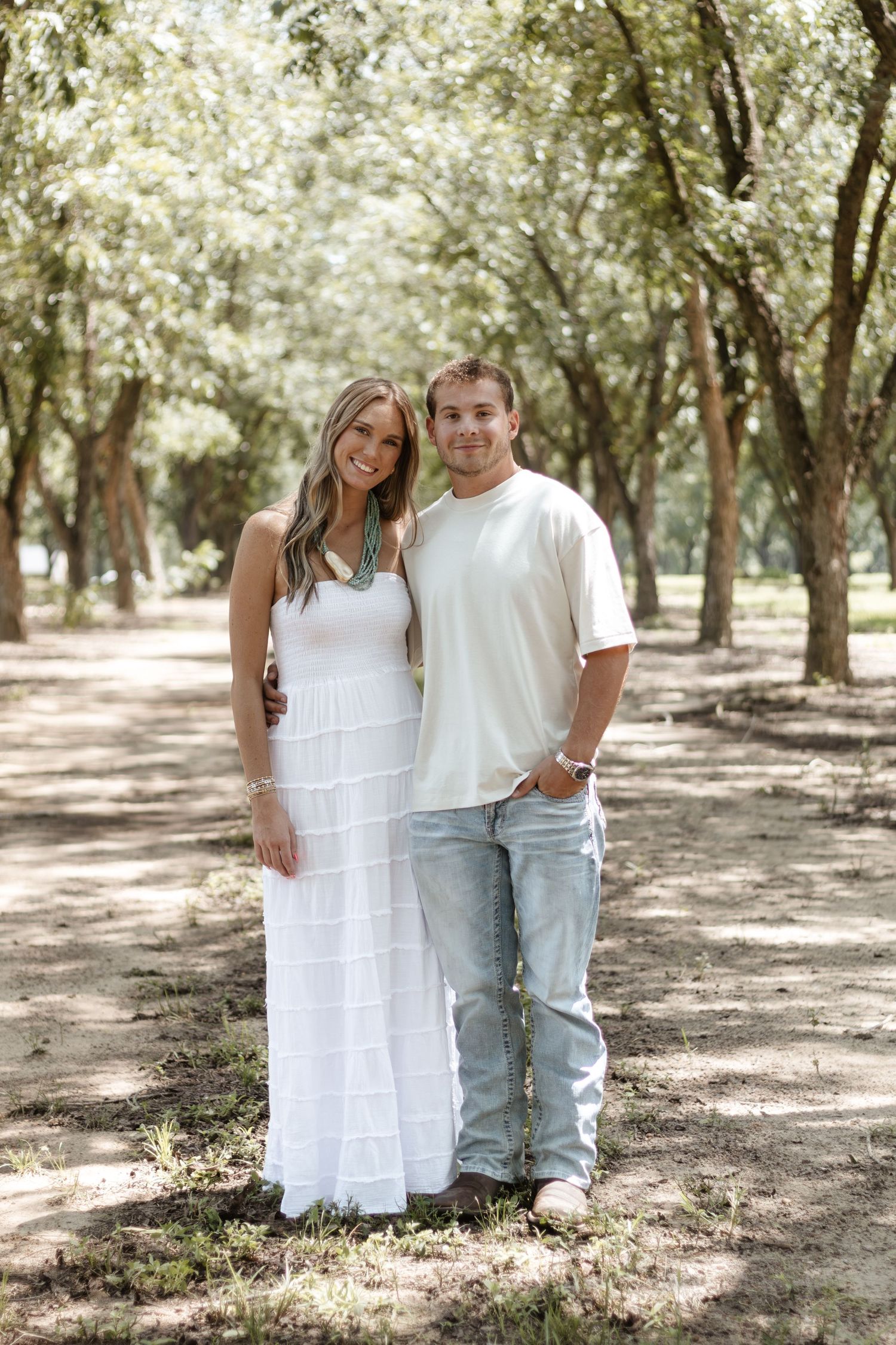 Smiling couple in white outfits posing together on a tree-lined path in dappled sunlight.