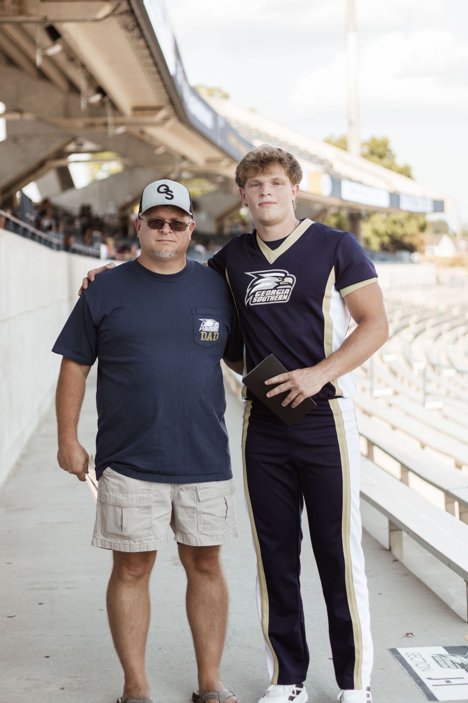 Father and son posing together in stadium stands, son wearing Georgia Southern football uniform.