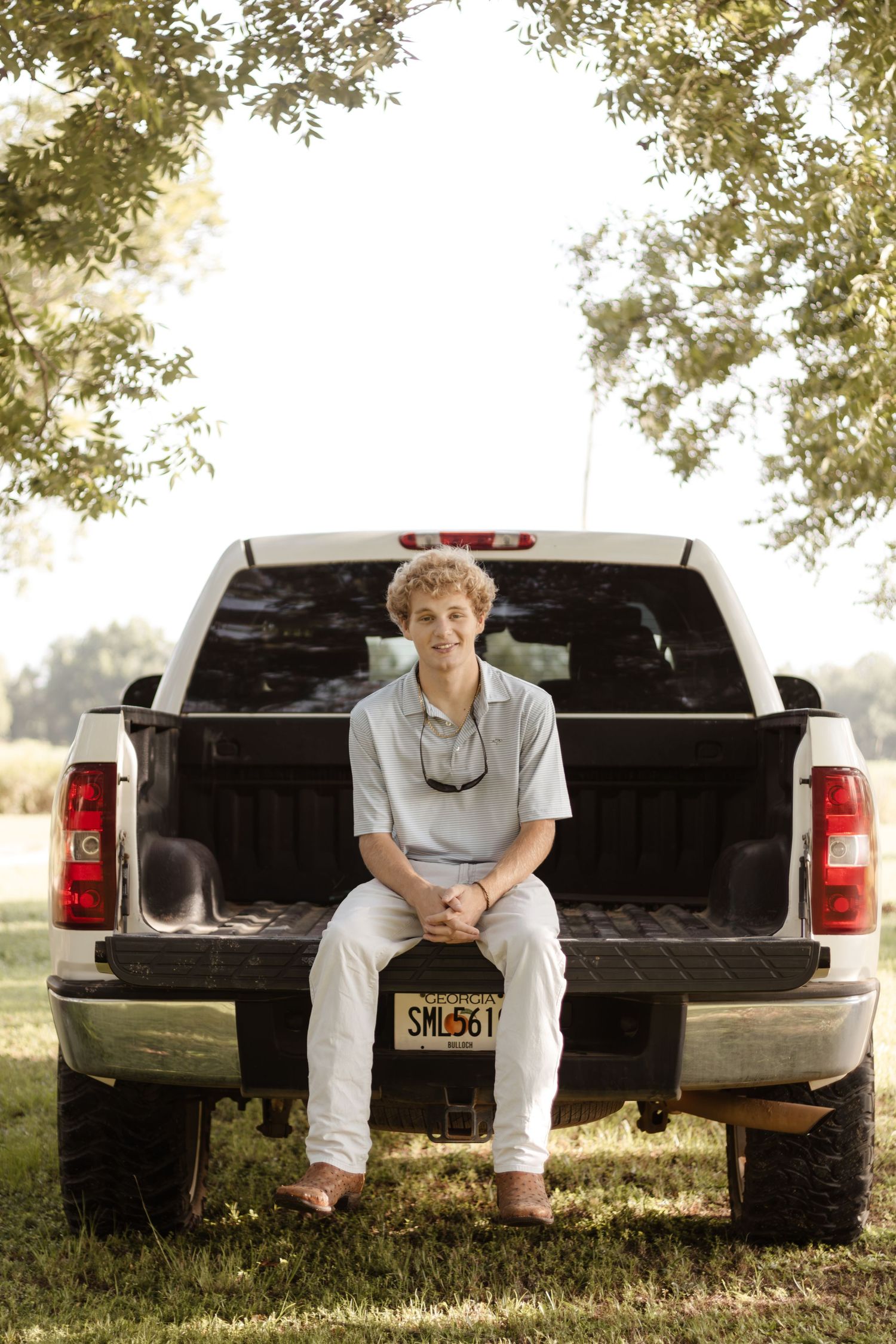 Smiling teenage boy with curly hair sitting on a truck tailgate under shaded trees outdoors.