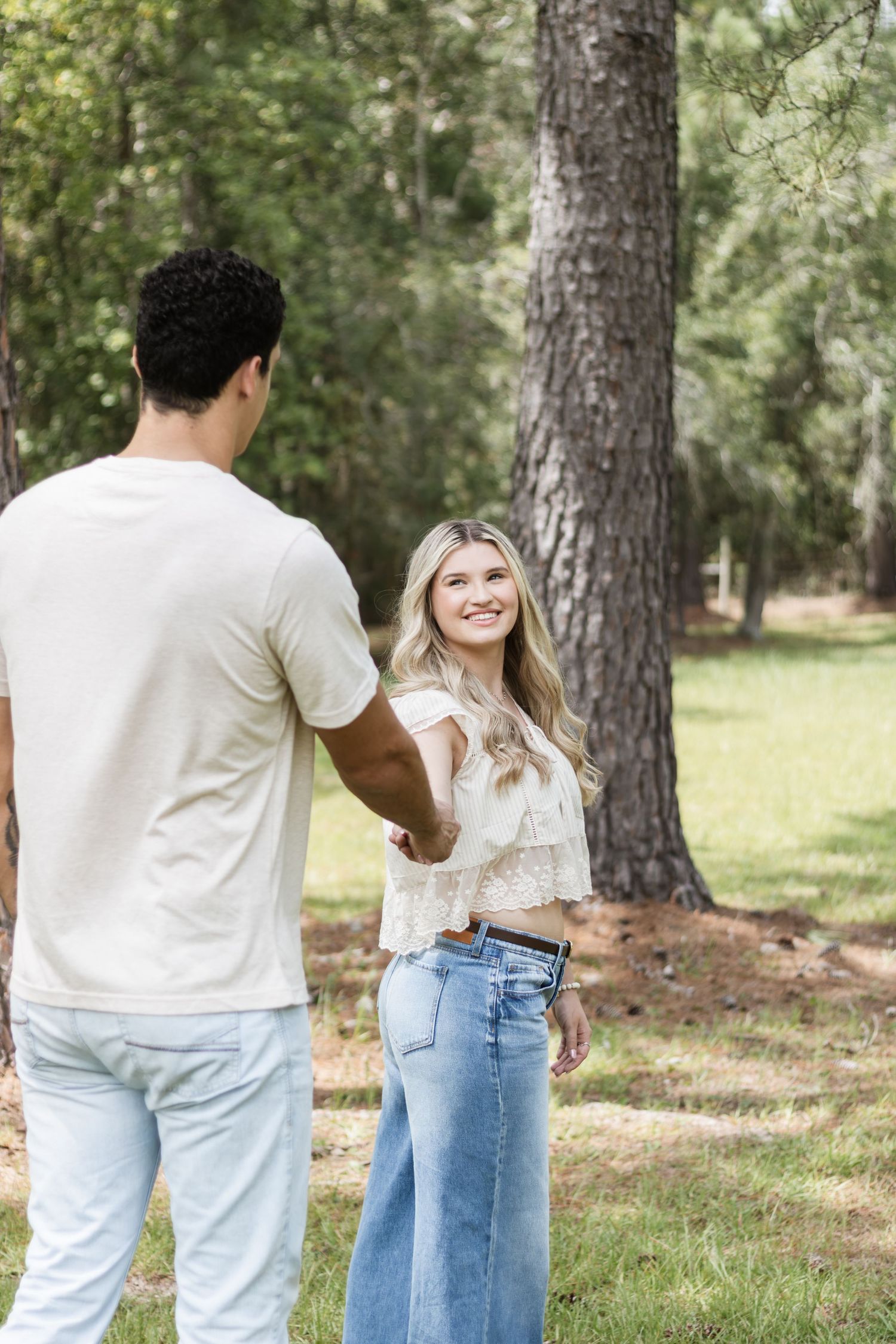 A smiling blonde woman in jeans holds hands with a man while standing among tall pine trees.