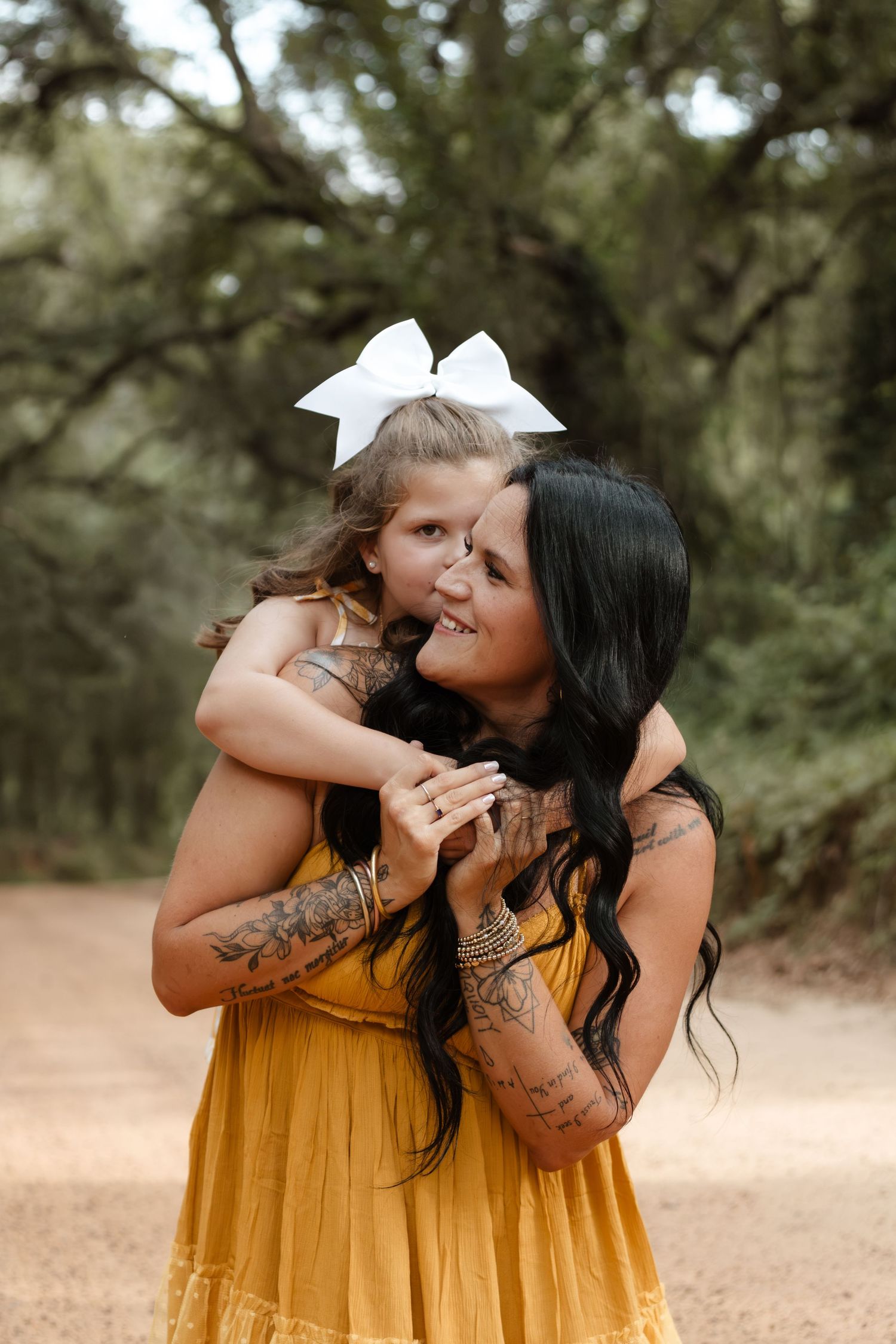 A tattooed woman in a yellow dress lovingly holds a young girl with a white hair bow outdoors.