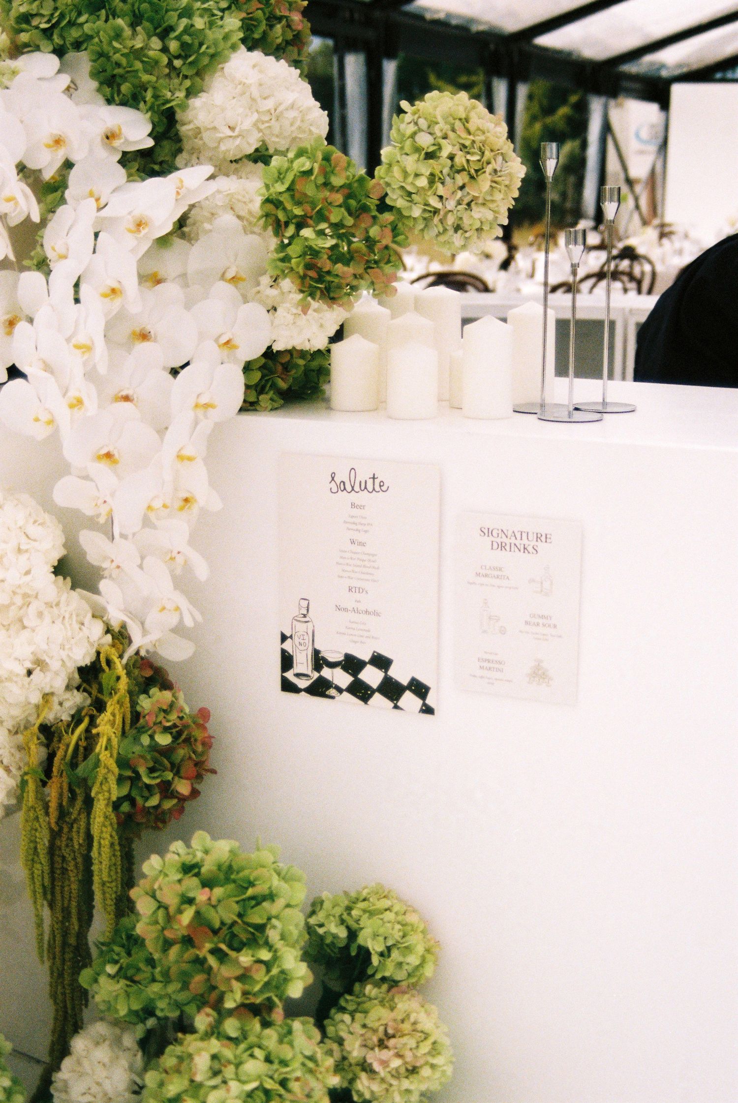 Elegant wedding bar decorated with white orchids and green hydrangeas displaying signature drinks menu cards.