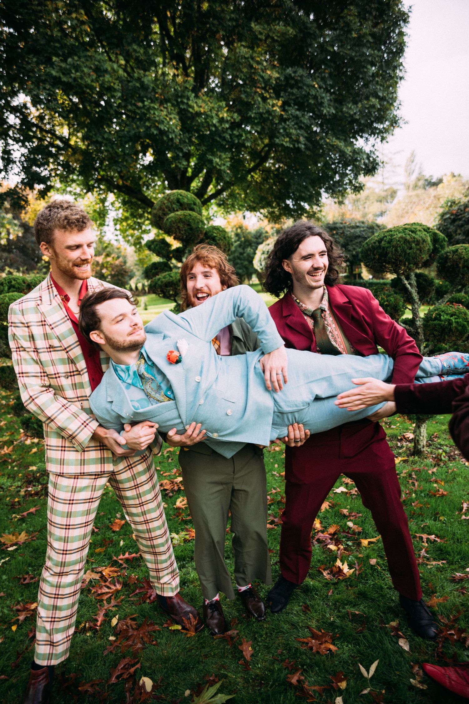 Laughing groomsmen in vibrant suits playfully lift the groom in a lush green garden.
