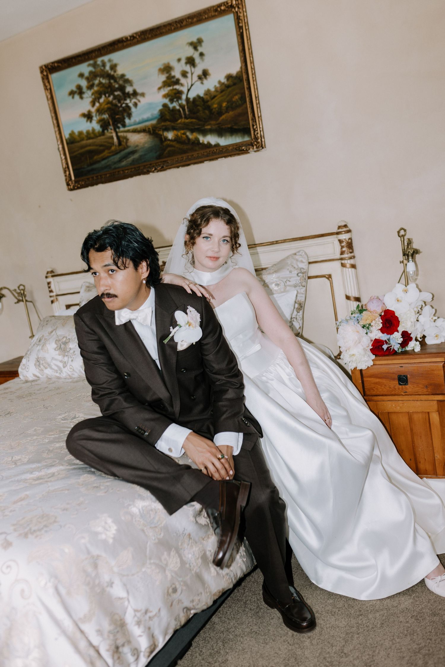 Bride and groom in vintage-style wedding attire seated on bed in classic bedroom with landscape painting above.