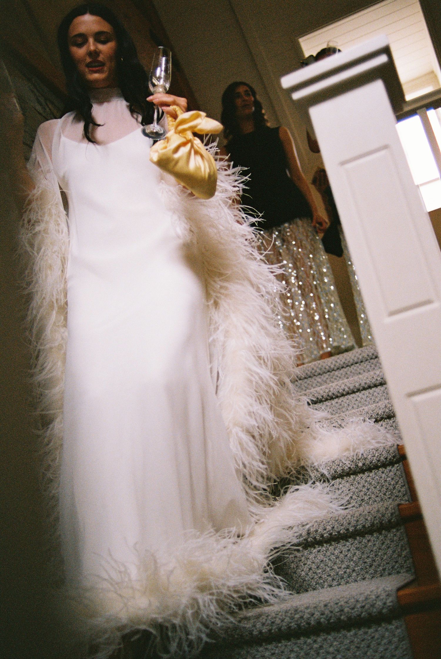 Bride in white feather-trimmed gown holds champagne glass on staircase before wedding.