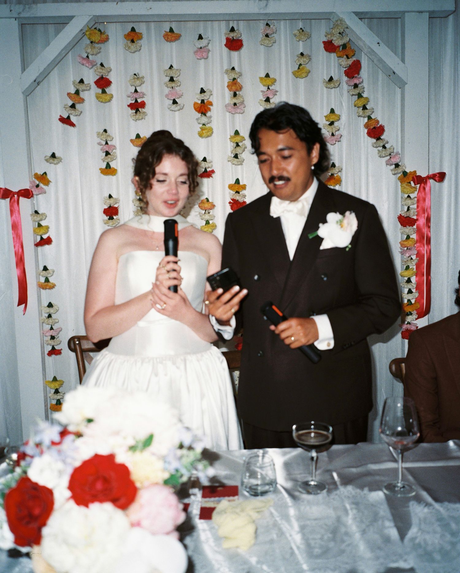 Bride and groom give speech at wedding reception with colorful paper garland backdrop.