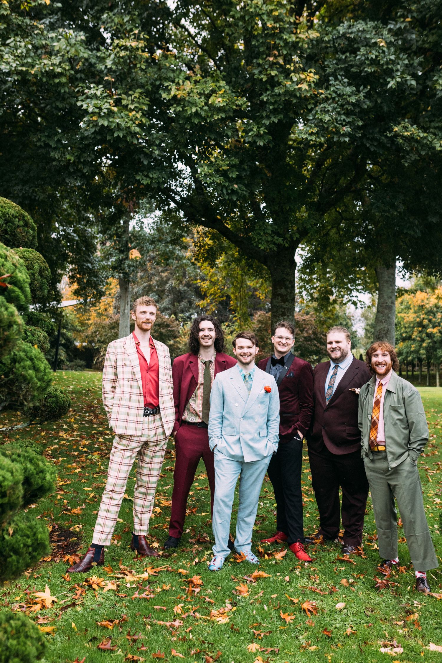 Six groomsmen in colorful eclectic suits pose together in an autumn garden with fallen leaves.