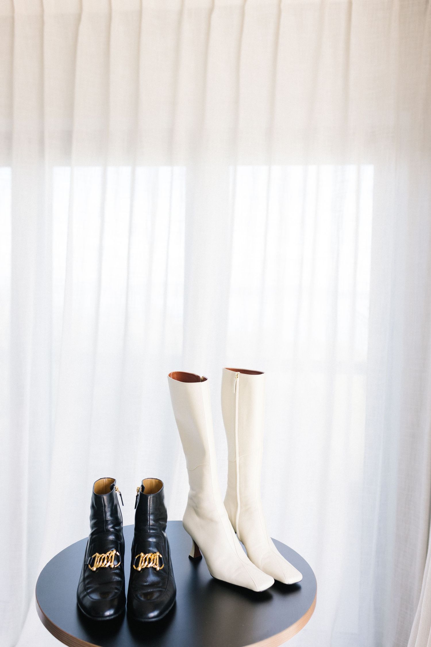 Black ankle boots and white knee-high heeled boots displayed on a round table against sheer curtains.