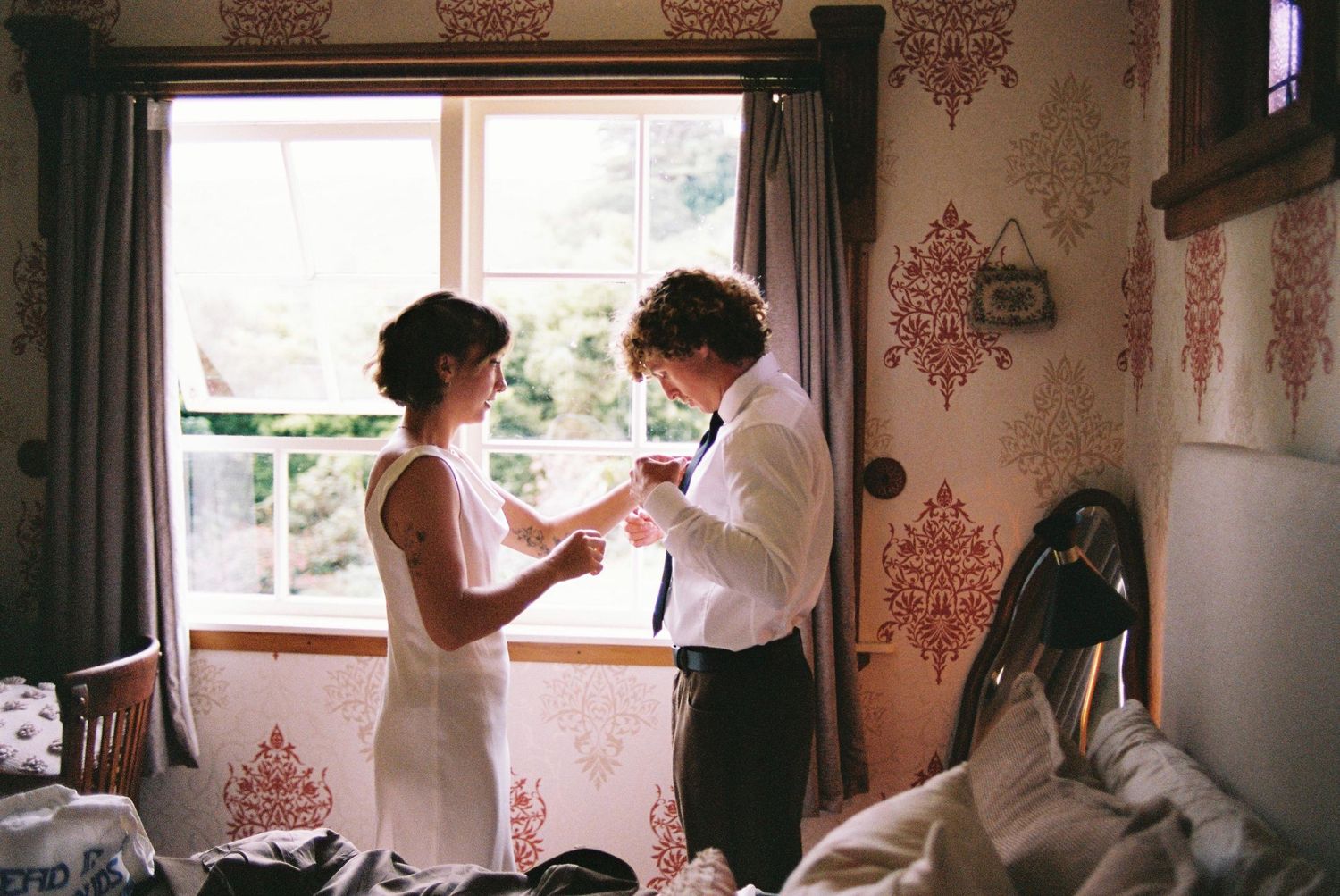 Bride and groom getting ready in a room with floral wallpaper and natural window light.