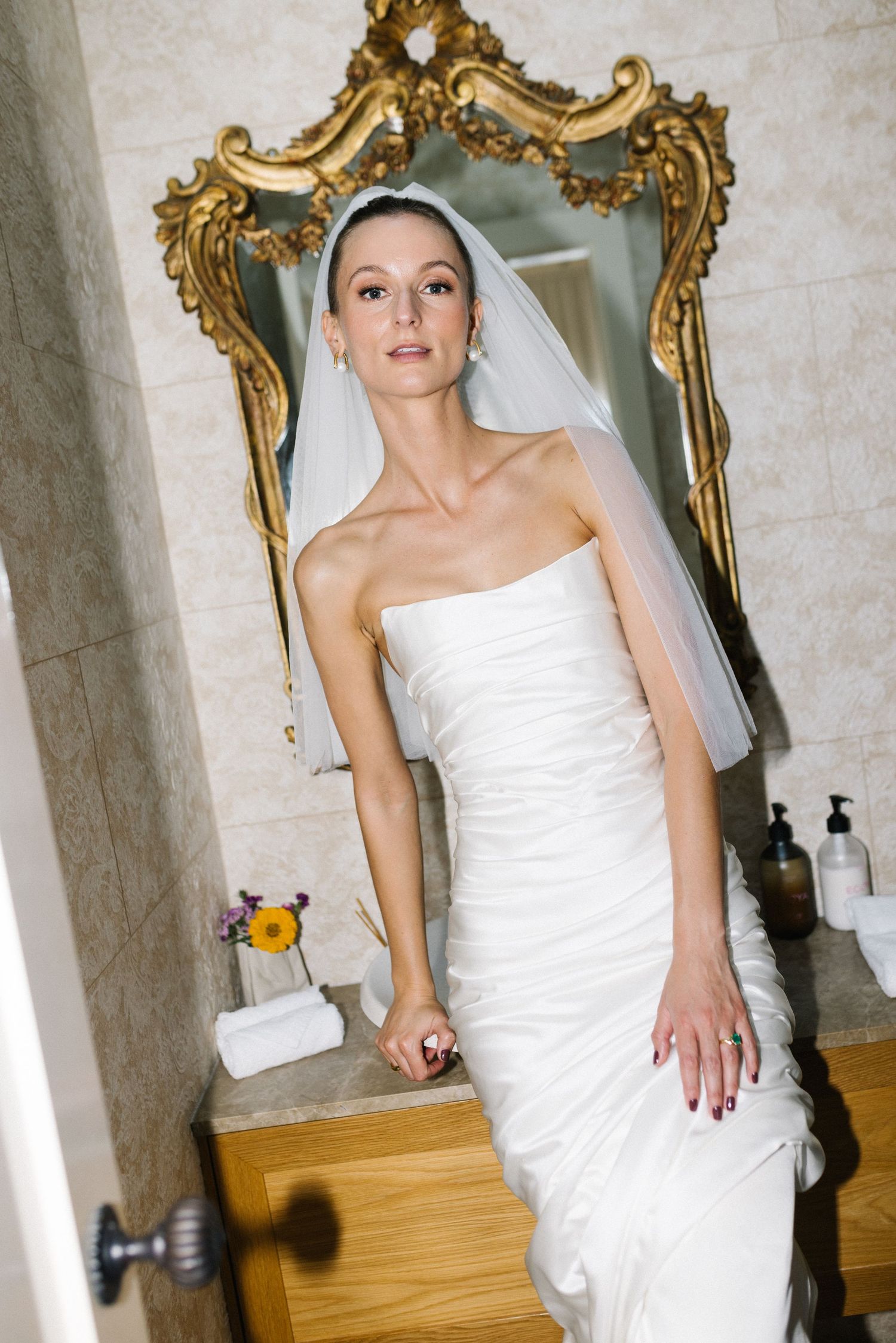 Elegant bride in strapless ruched white gown and veil posing before ornate gold-framed mirror in bathroom.