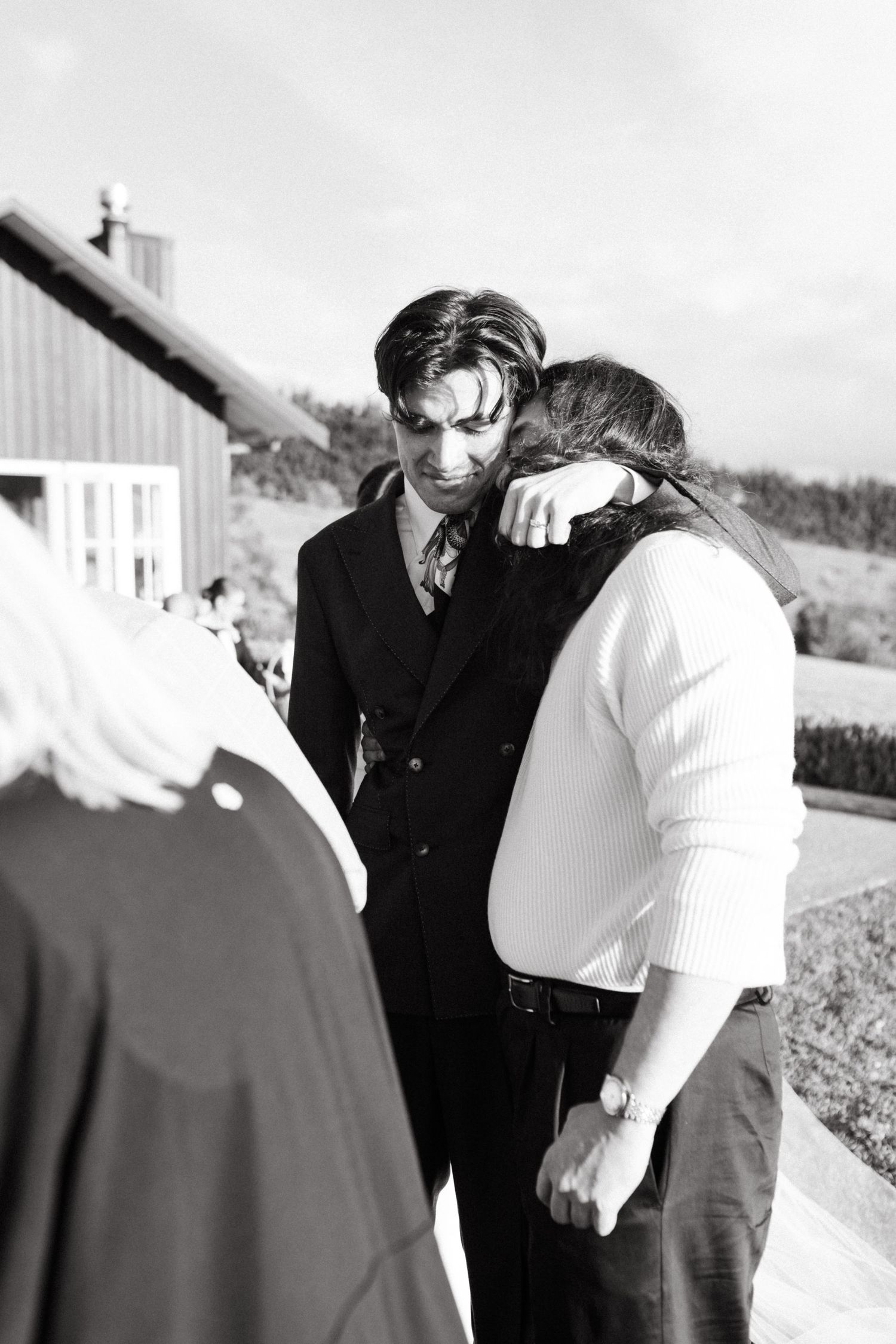 Black and white photo of groom in black suit comforting a guest outdoors at a wedding reception.