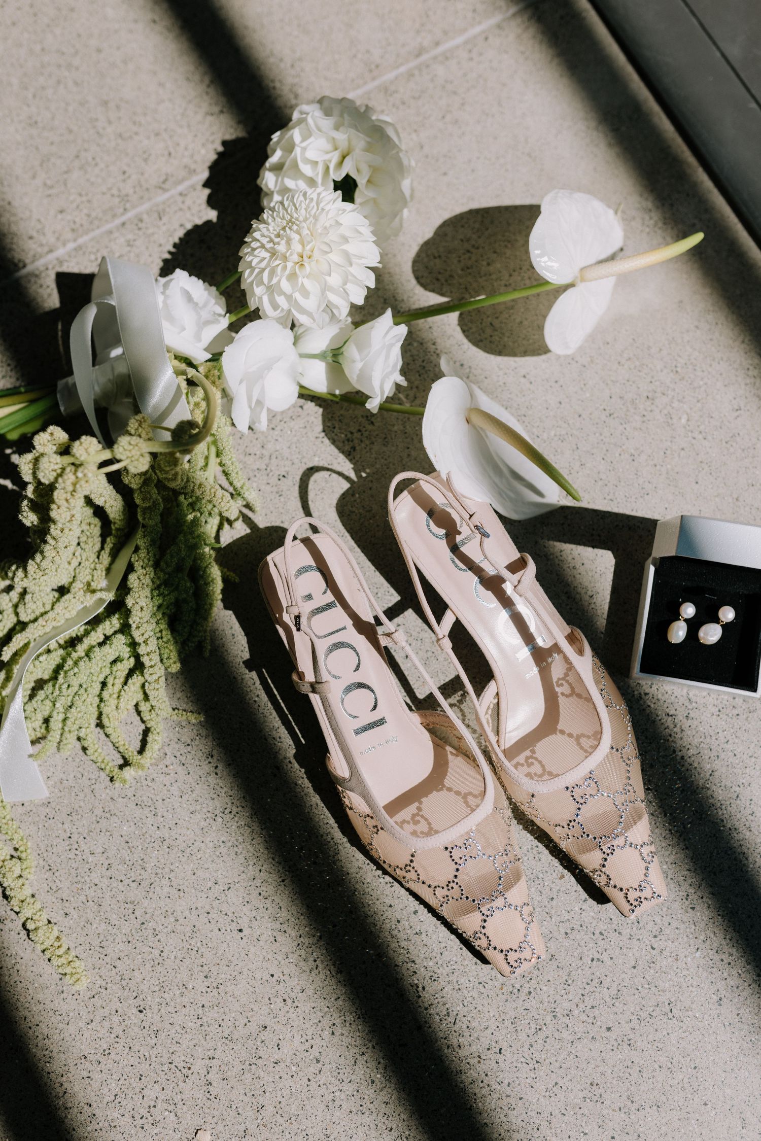 Gucci slingback heels beside white wedding bouquet and pearl earrings on textured floor.