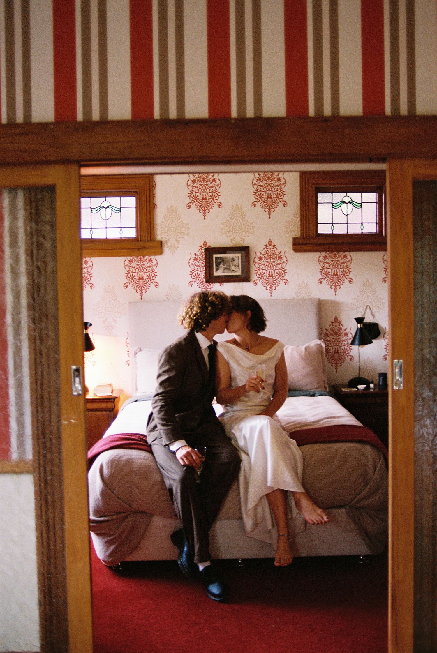 Newlyweds kissing on bed in vintage-styled room with striped walls and stained glass windows.