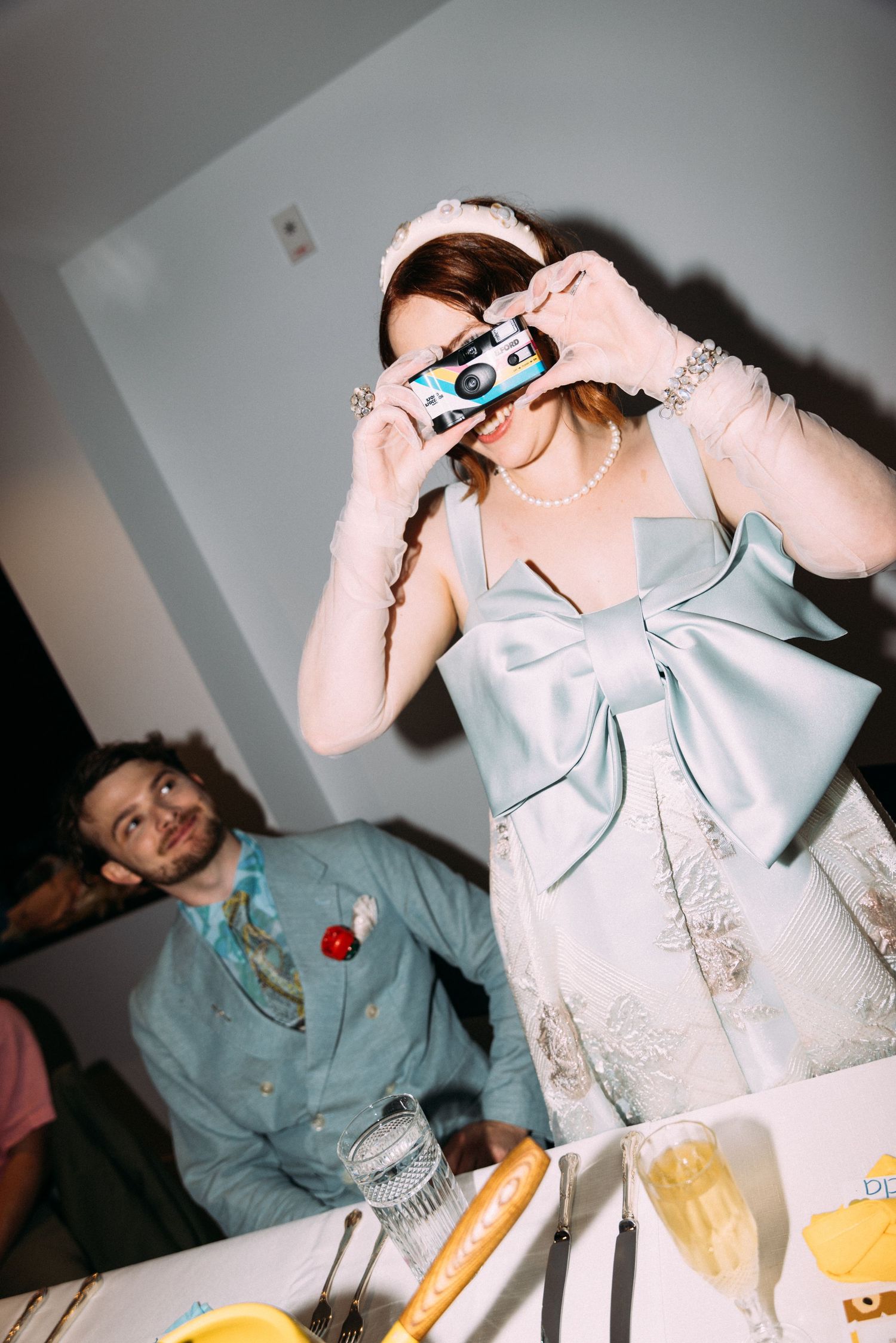 Bride in light blue bow gown photographs guests with a disposable camera at wedding reception.