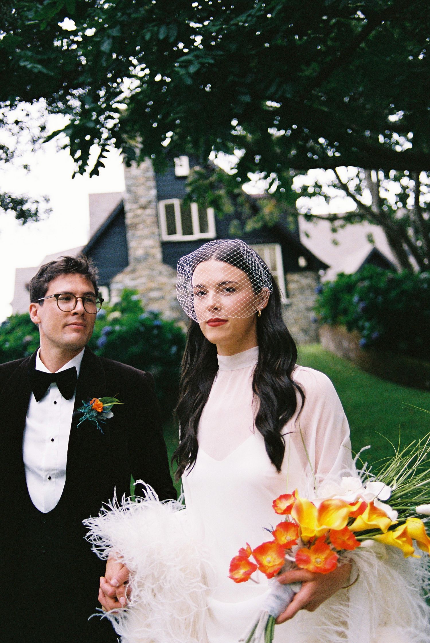 Bride in feathered gown with birdcage veil holds orange bouquet beside groom in tuxedo.
