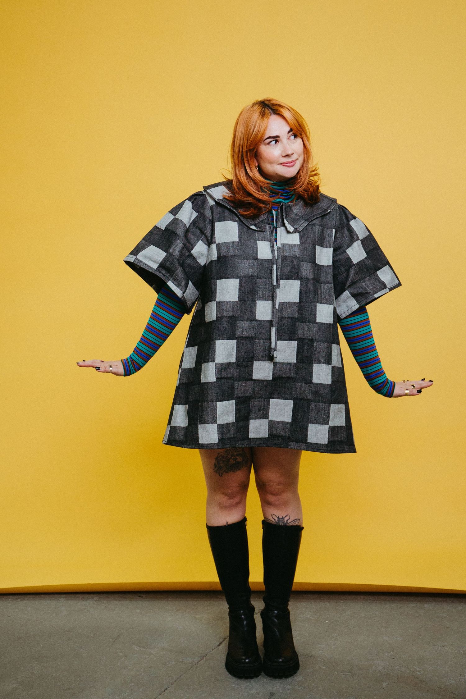 Woman with red hair wearing checkered poncho dress posing against a yellow background.
