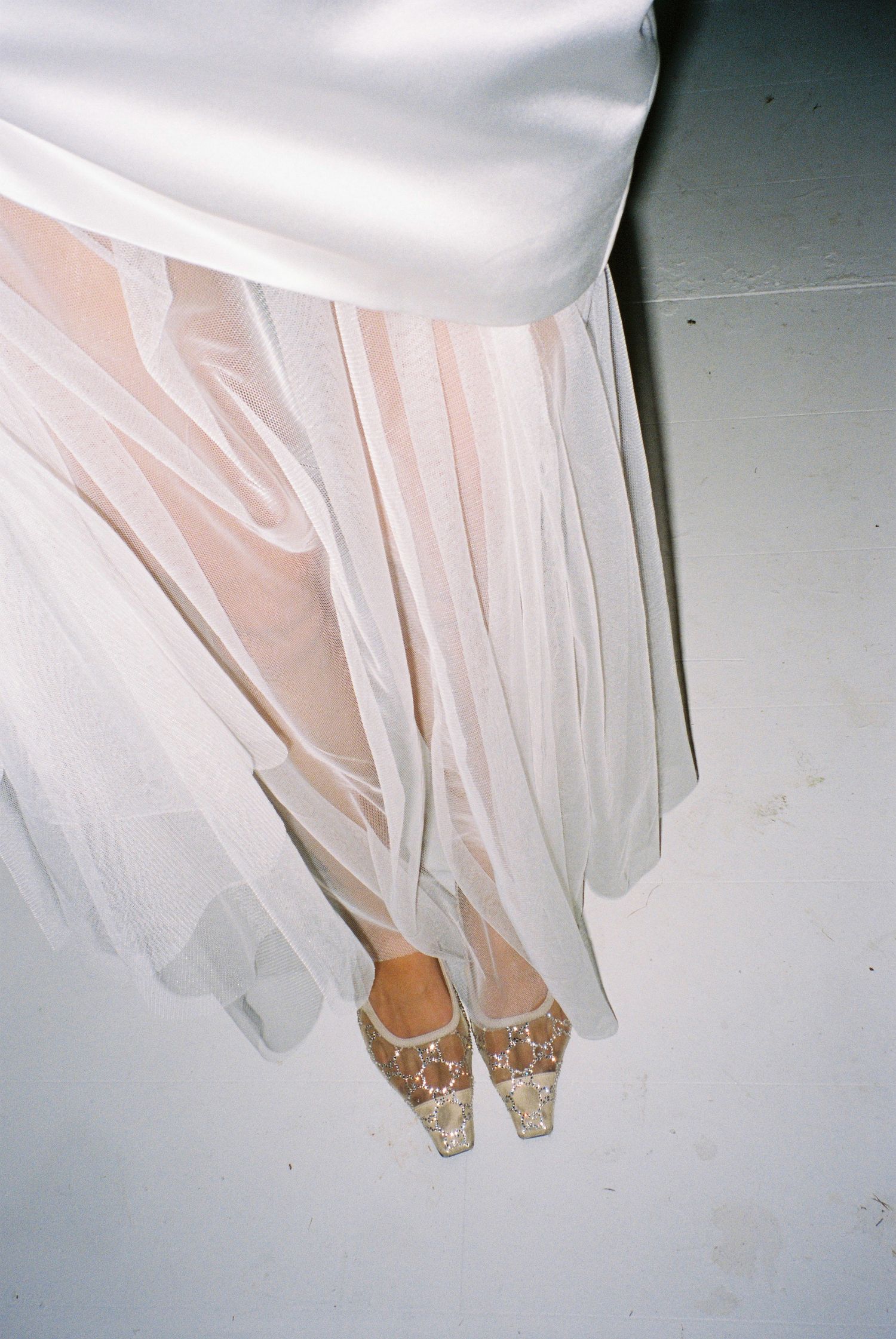 Close-up of bride's sheer white tulle skirt and embellished gold heels on white floor.
