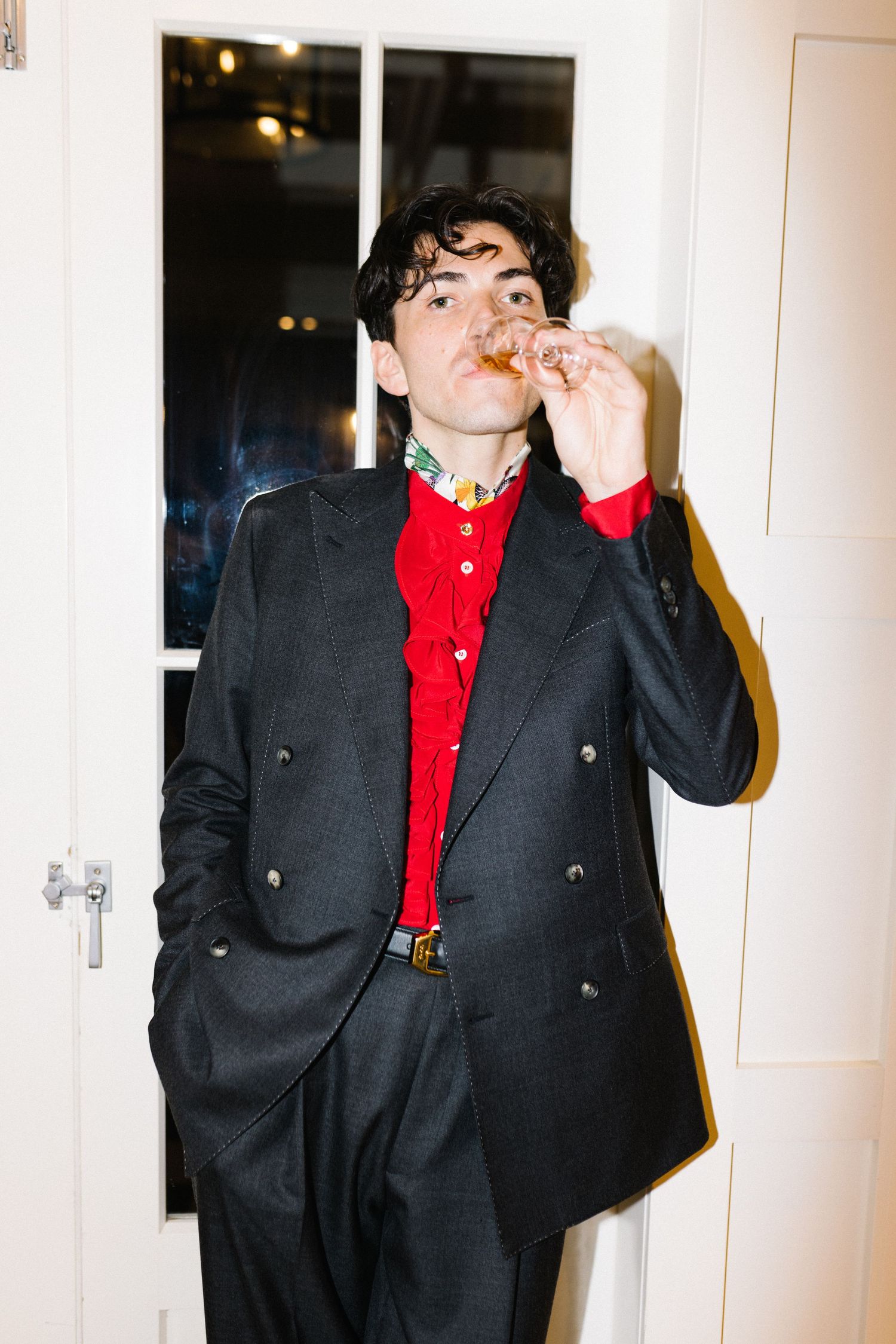Man in black double-breasted suit and red ruffled shirt poses indoors at a wedding reception.