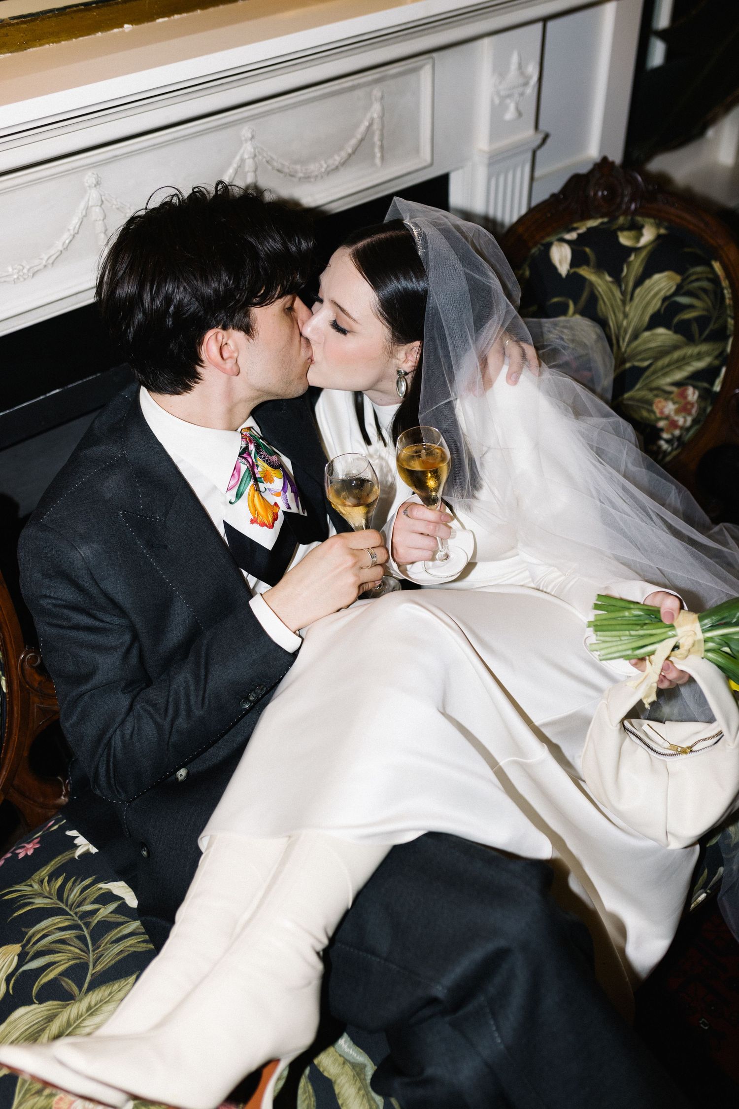Bride and groom share a kiss while holding champagne glasses seated on a floral chair by a fireplace.
