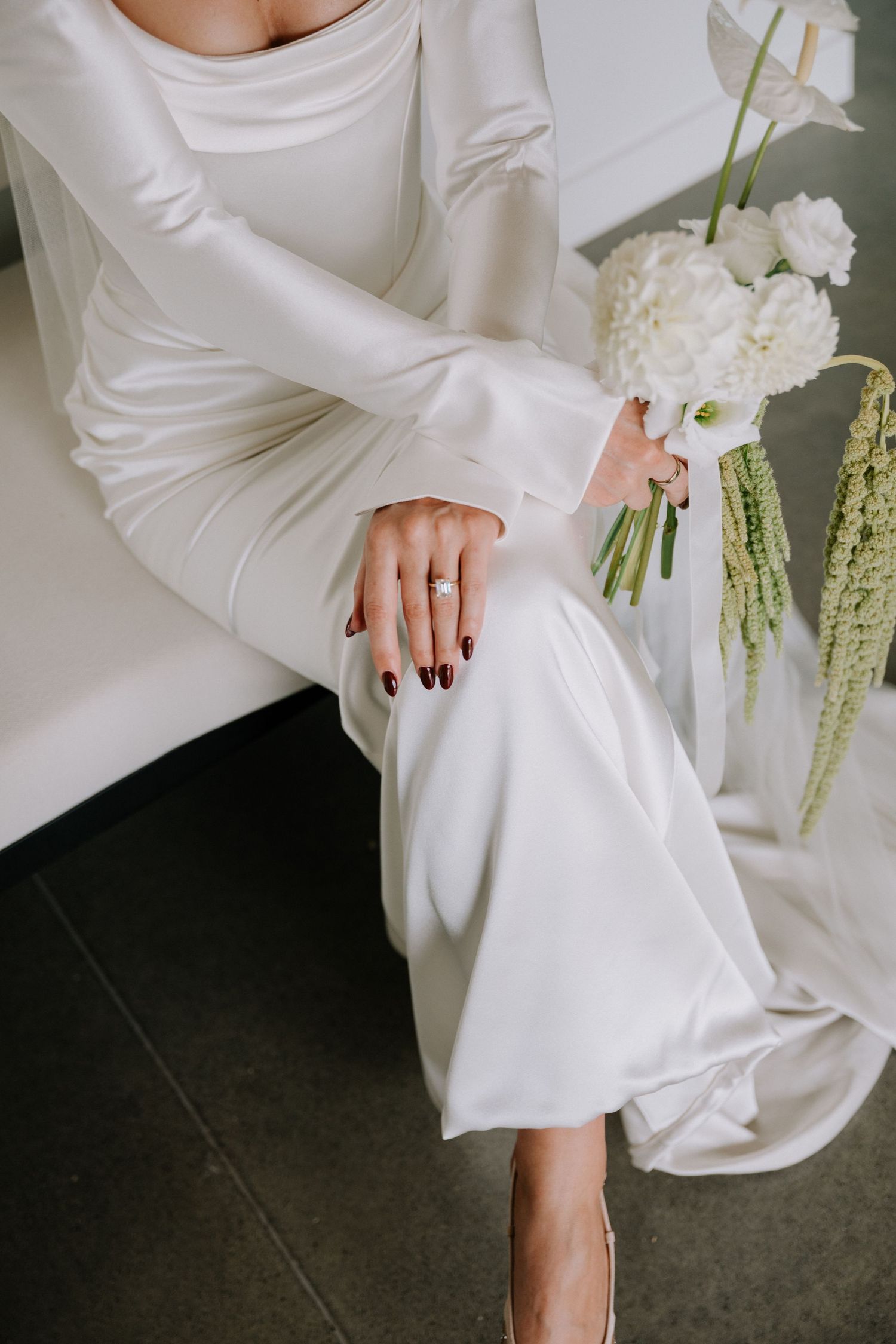 Bride in white satin gown with dark red nails and diamond ring holding white floral bouquet.