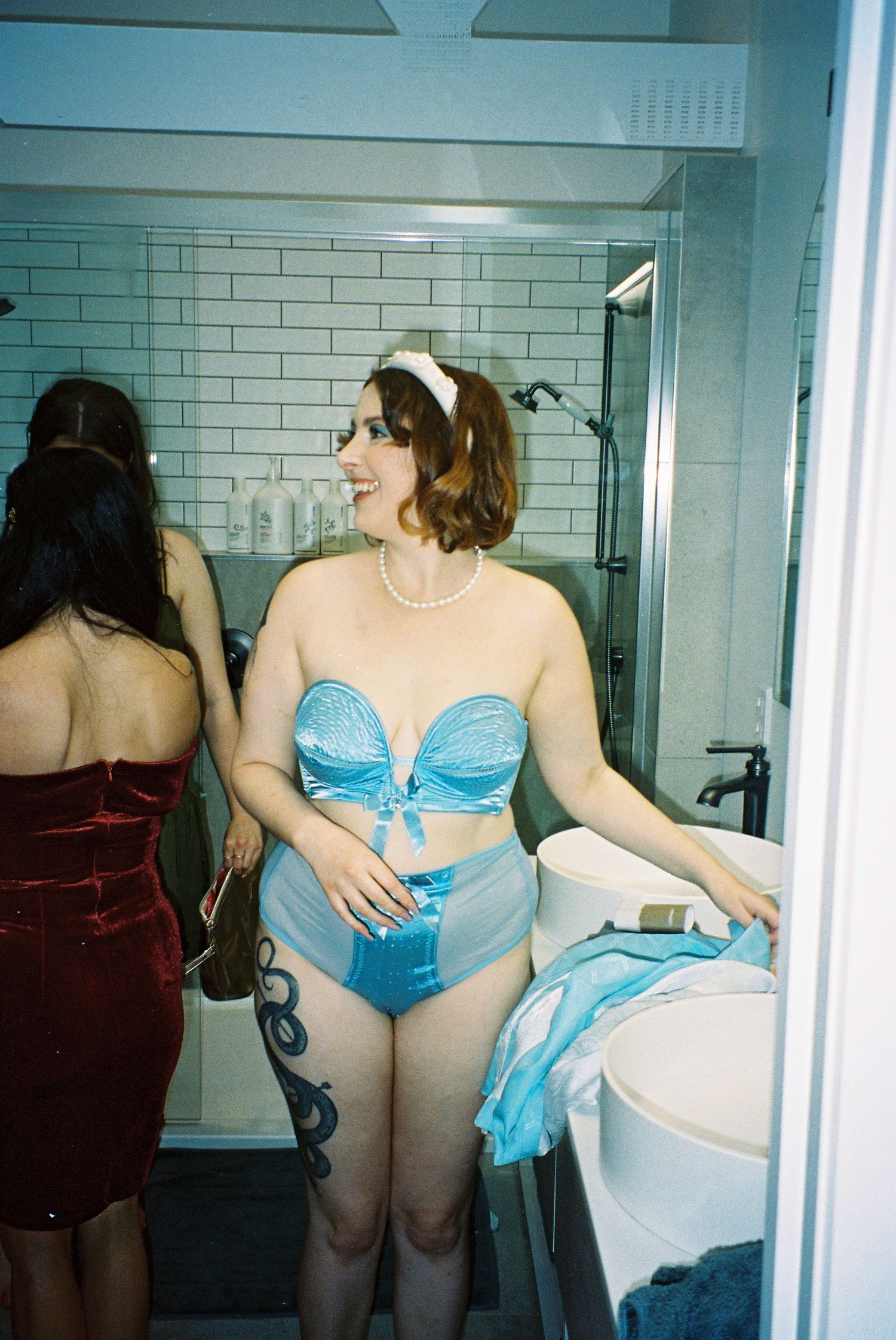 Woman in blue vintage-style lingerie set with pearl necklace stands in white tiled bathroom.